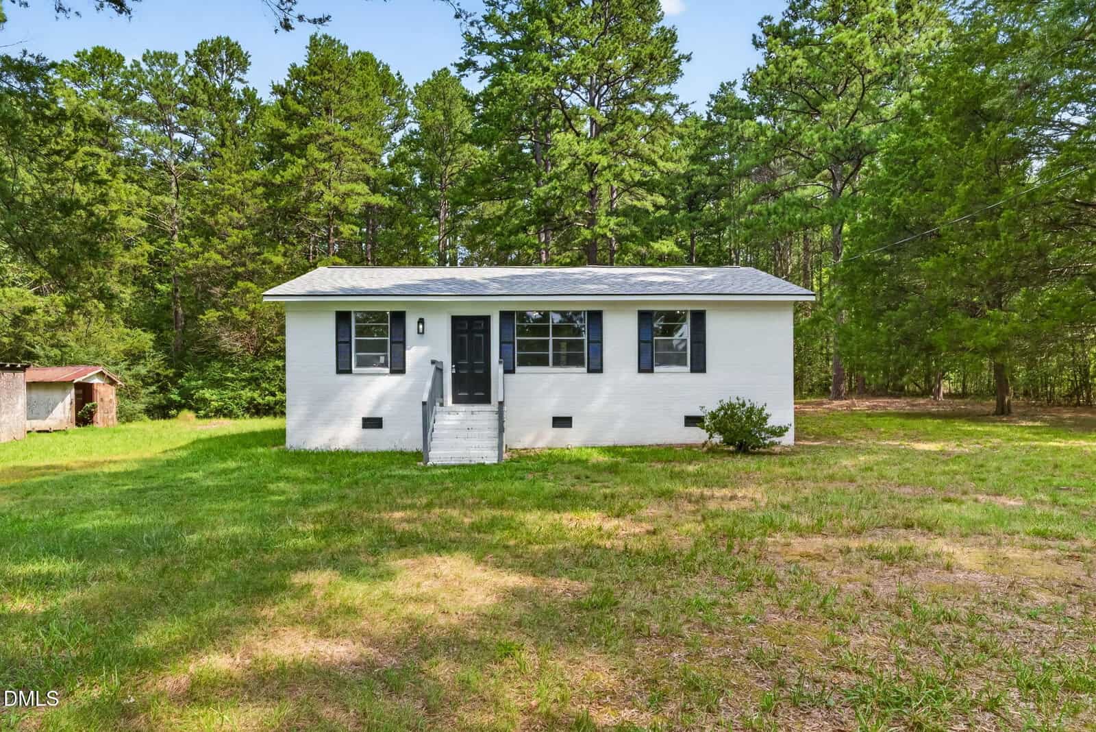 Small white brick house with black shutters and door, set in a grassy yard surrounded by tall trees. A few steps lead to the front door, and there’s a small shrub near the right corner of the house.