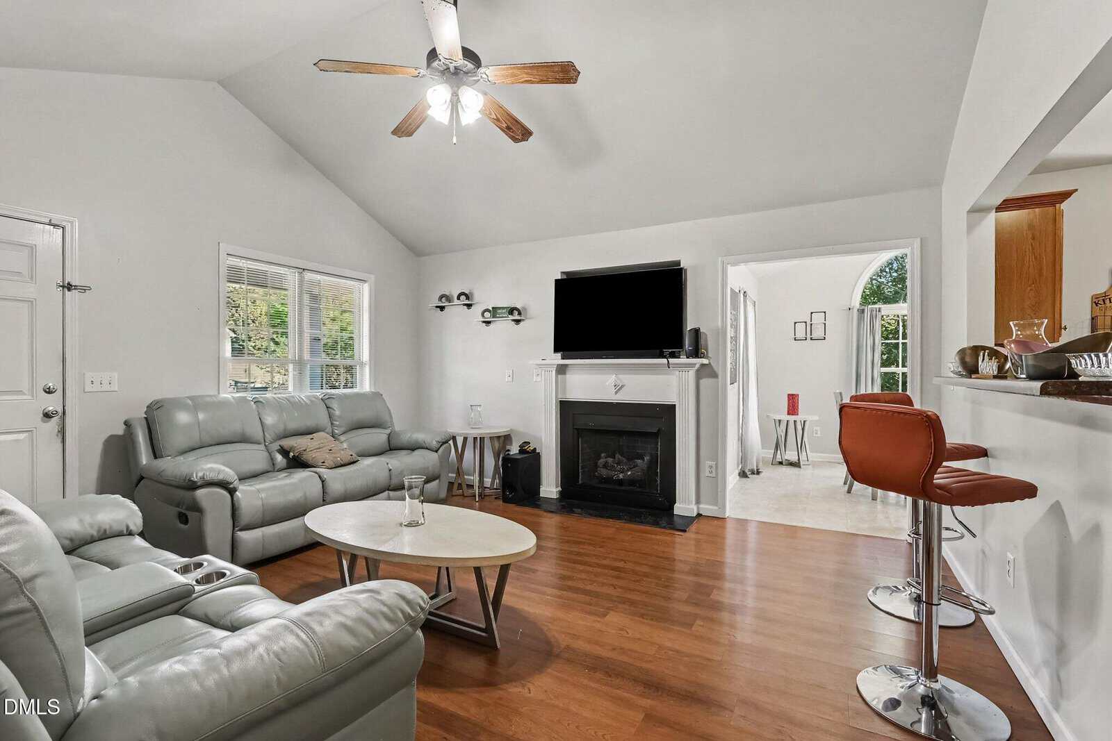 Modern living room with gray leather sofas, a round coffee table, hardwood floors, a ceiling fan, a fireplace with a TV above, and a breakfast bar with orange stools, opening to a bright dining area.