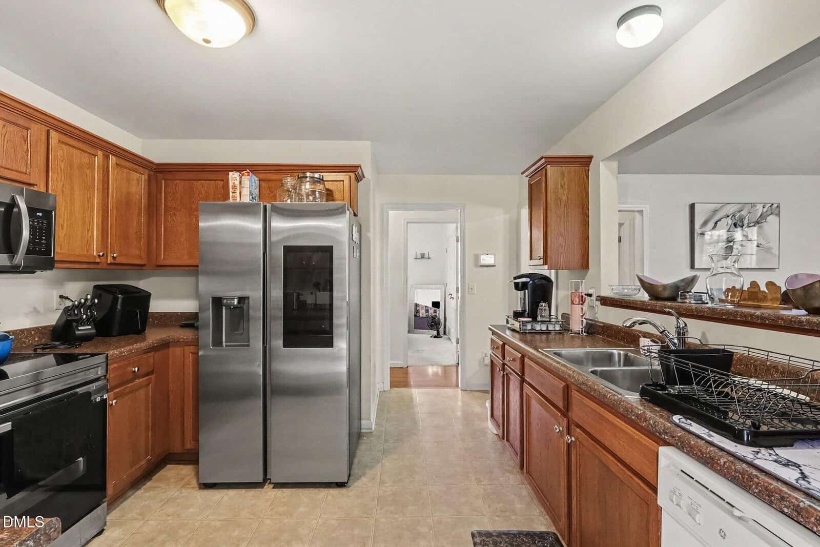 Modern kitchen with stainless steel appliances, wooden cabinets, double sink, dish rack, and coffee maker. A hallway leads to another room in the background. Countertops have various kitchen items.
