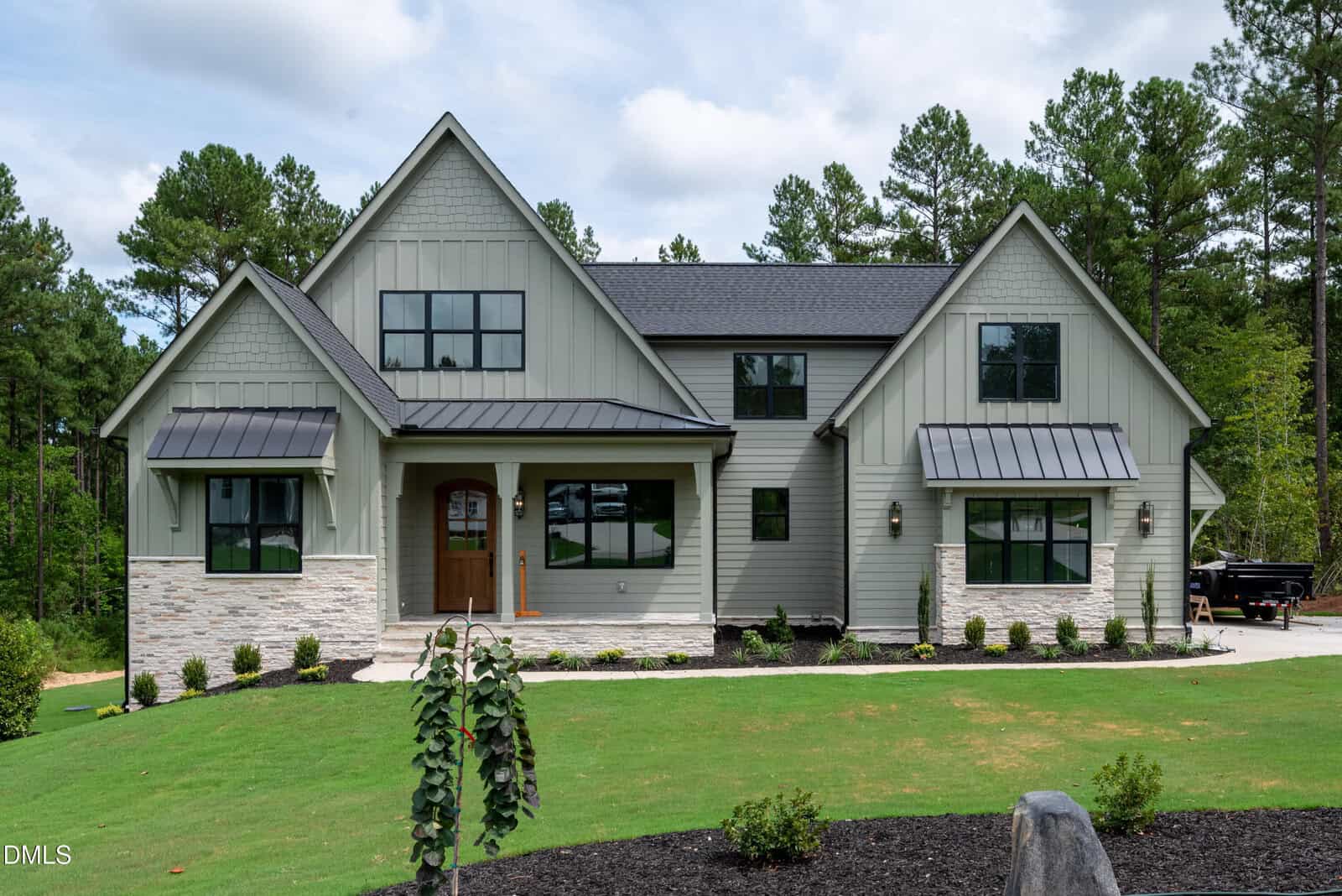 A modern two-story house at 230 Whistlers Cove, Louisburg, with gray siding, black trim, and a stone facade sits on a green lawn with landscaped bushes and trees. The front door is wooden with glass panes. The sky is partly cloudy.