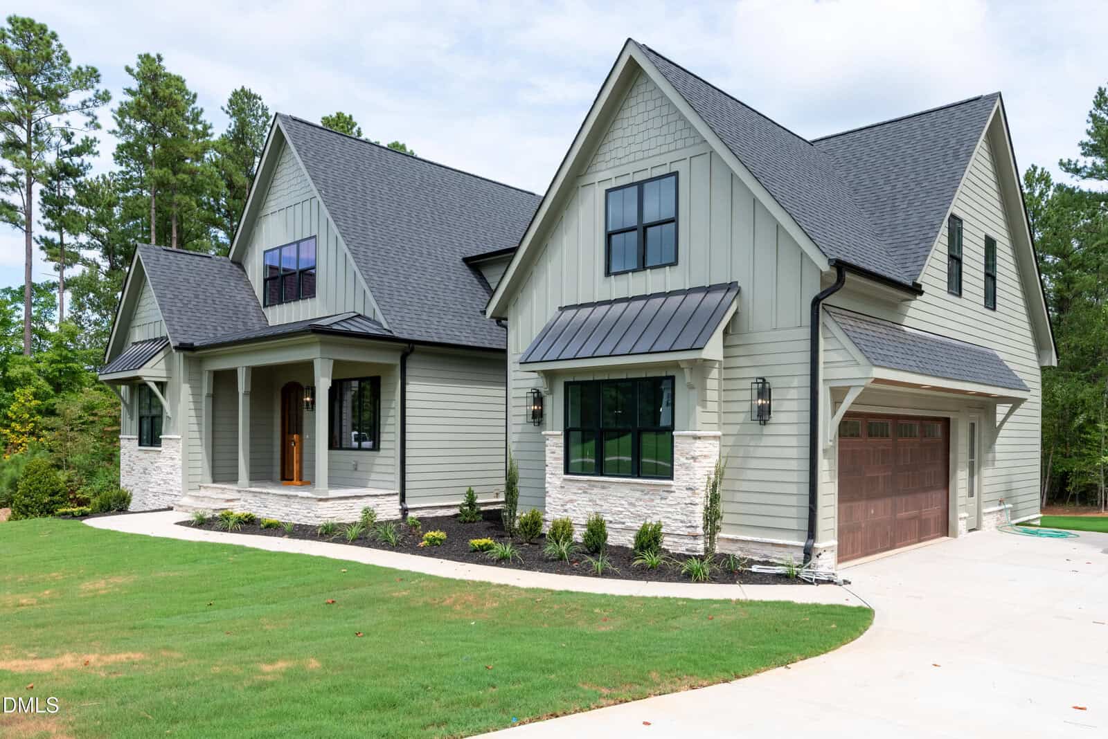 Modern two-story house with grey siding, stone accents, black-framed windows, and a brown double garage. Neatly landscaped front yard and concrete driveway with trees in the background.