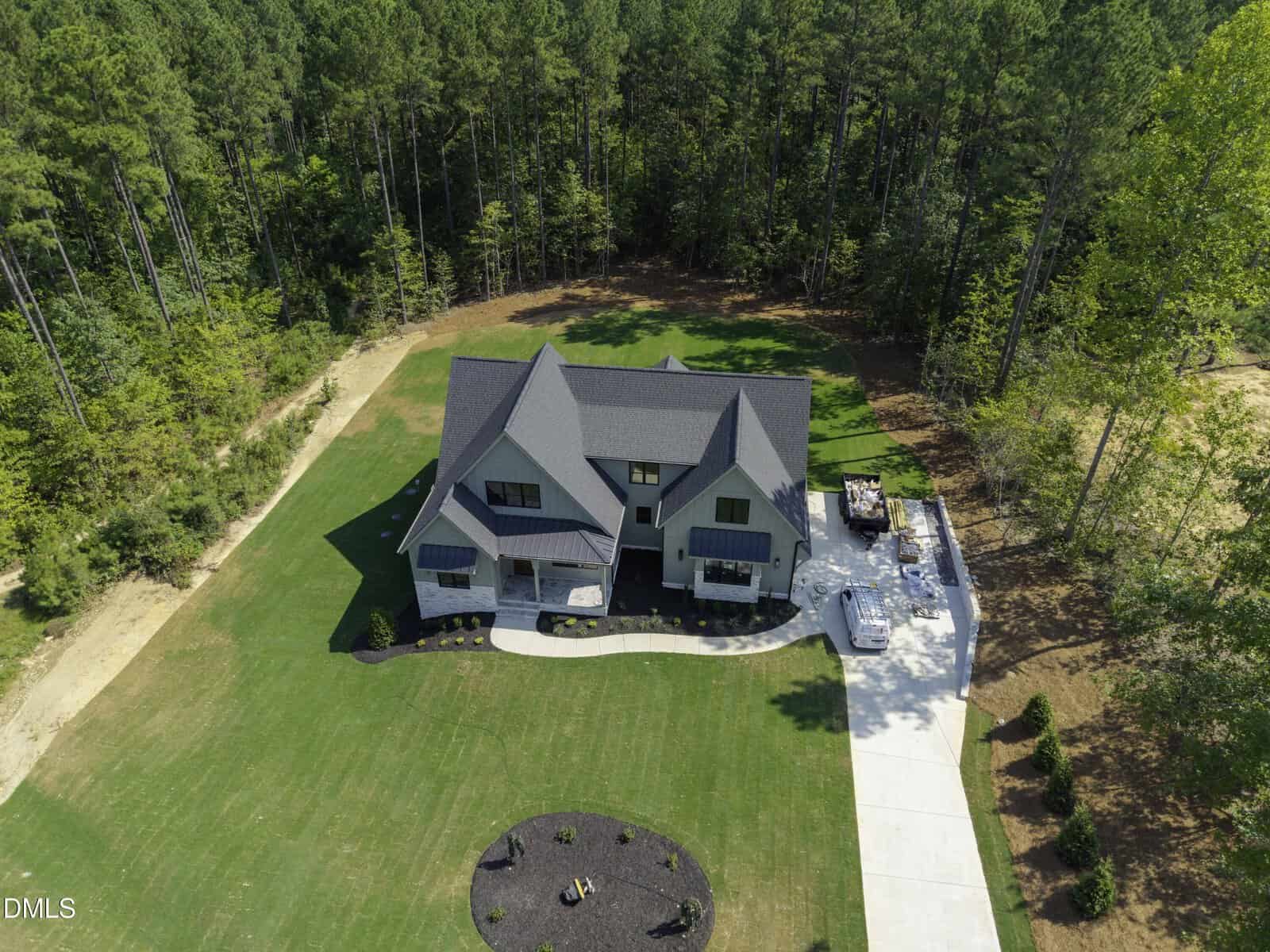A large modern house with a dark roof, set in a spacious green yard surrounded by trees, features a circular fire pit area, a long driveway, and a backyard patio with outdoor furniture.