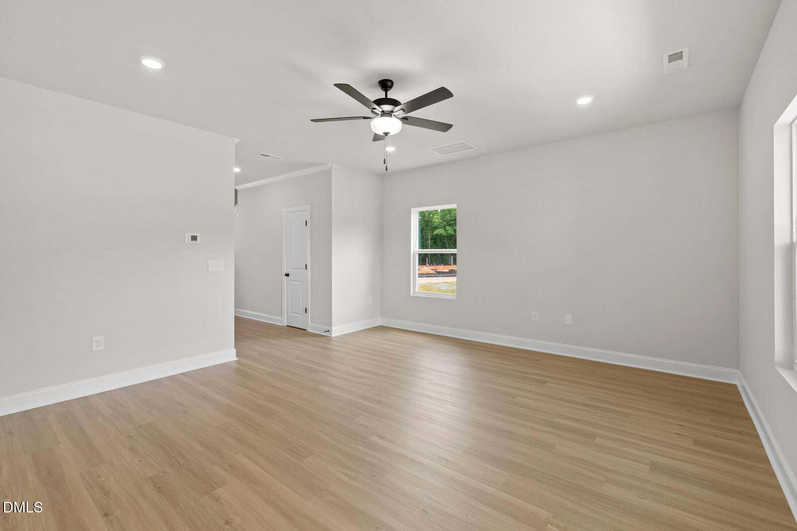 Bright, empty living room with light wood floors, white walls, ceiling fan with lights, recessed lighting, and two windows. A door and hallway are visible on the left side.
