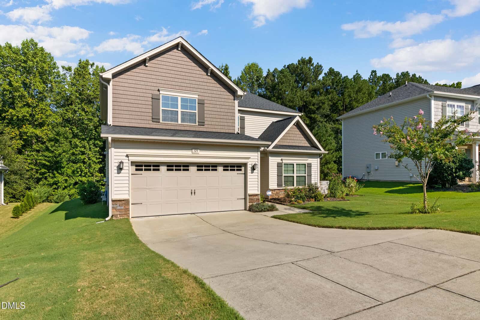 A two-story suburban house with a double garage, beige siding, and brown accents. It has a curved driveway, green lawn, small shrubs, and trees in the background under a partly cloudy sky.