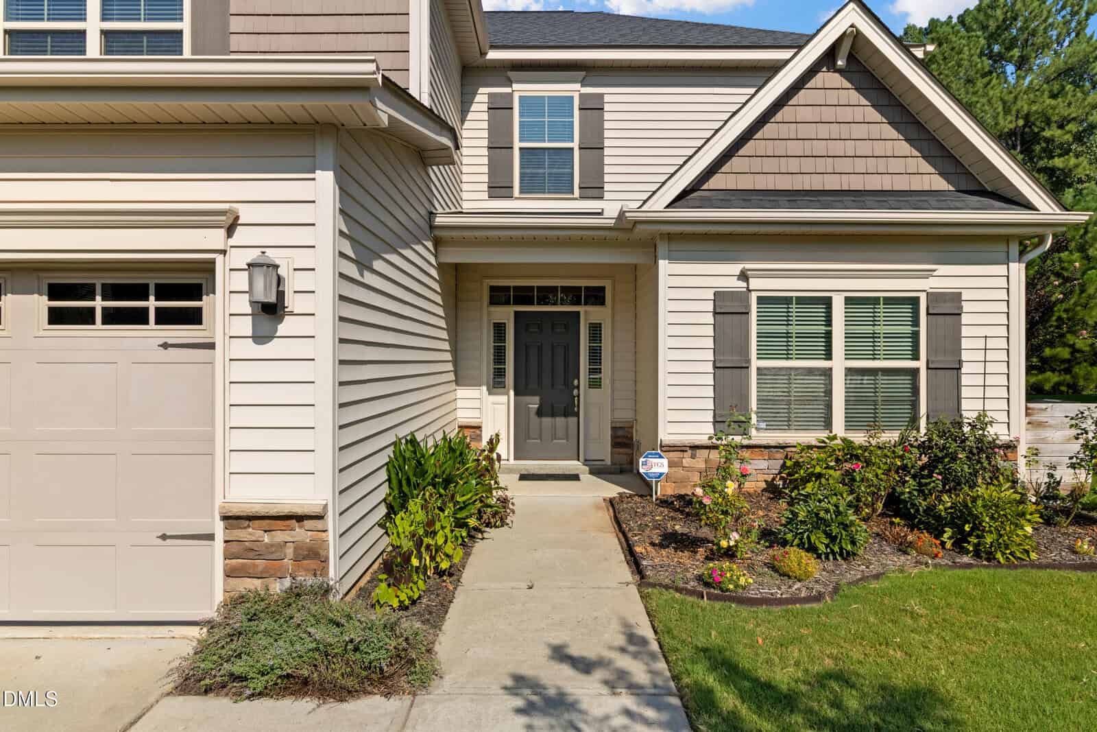 Front view of a beige house with a double front door, garage, white trim, shuttered windows, and landscaped garden with bushes and flowers along the sidewalk.