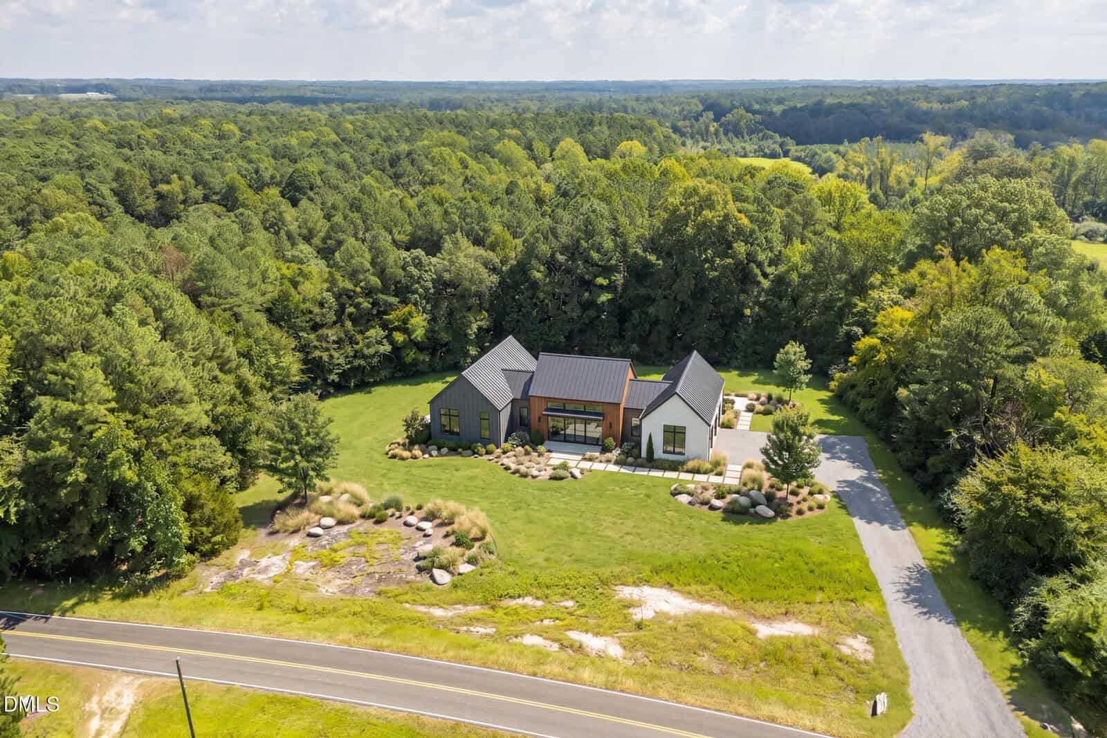 A modern single-story house with a dark roof sits in a large green yard surrounded by trees, with a driveway and a road in the foreground.