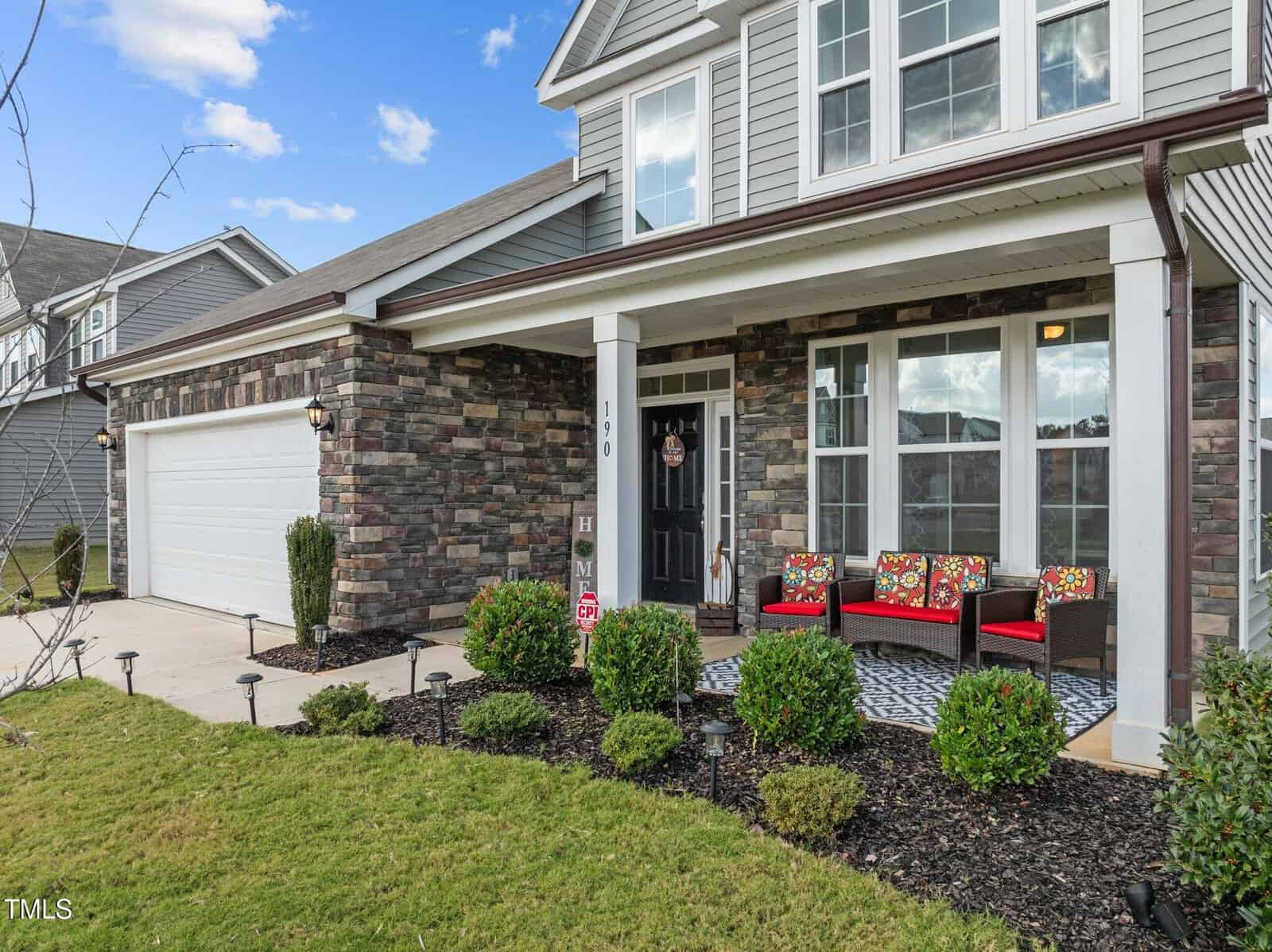 Charming two-story suburban house in Franklinton with a stone and siding exterior, attached garage, and inviting front porch. The porch at 190 Hickory Run Lane features red-cushioned chairs and a rug. Green shrubs and a manicured lawn are visible in the foreground under a blue sky.