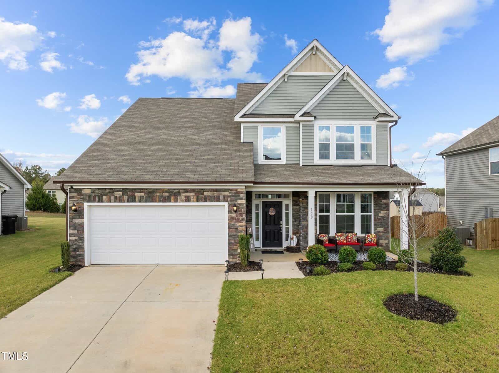 Two-story suburban house with gray siding, stone trim, and a white garage door. Well-kept lawn, small shrubs, and flower boxes decorate the front. The sky is blue with scattered clouds.