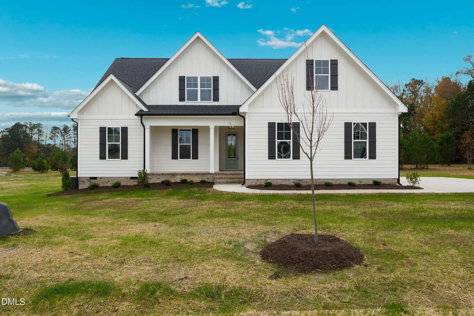 A white, single-story house with black shutters, a gray front door, gable roof, and a small front porch. The house sits on a large grassy yard with a young tree in the foreground and trees in the background.