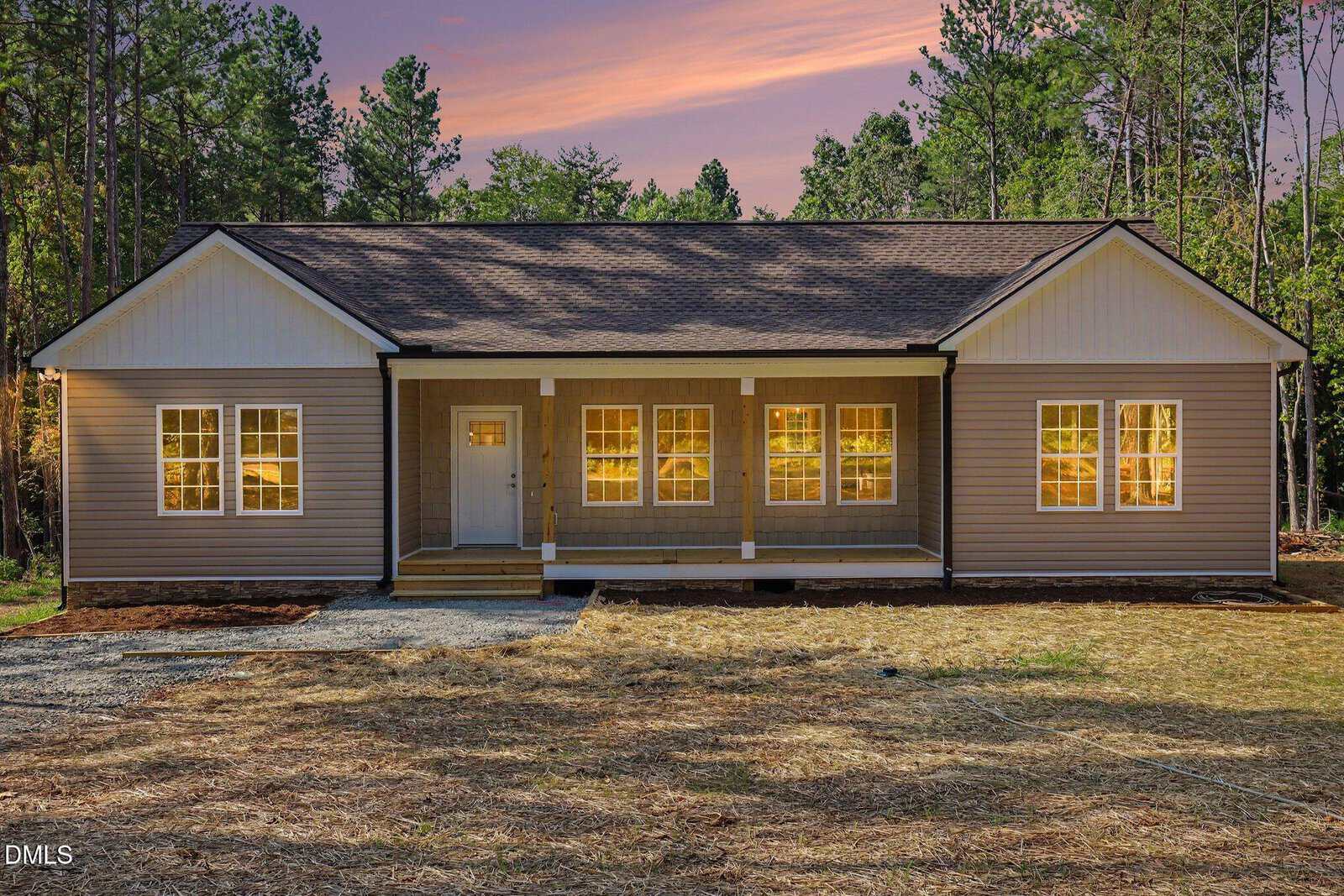 Single-story house with gray siding, white trim, a large front porch, and six lit windows on the front. Located at 2081 Satterwhite Road in Oxford, this home is surrounded by trees, with a dirt yard and a sunset sky in the background.