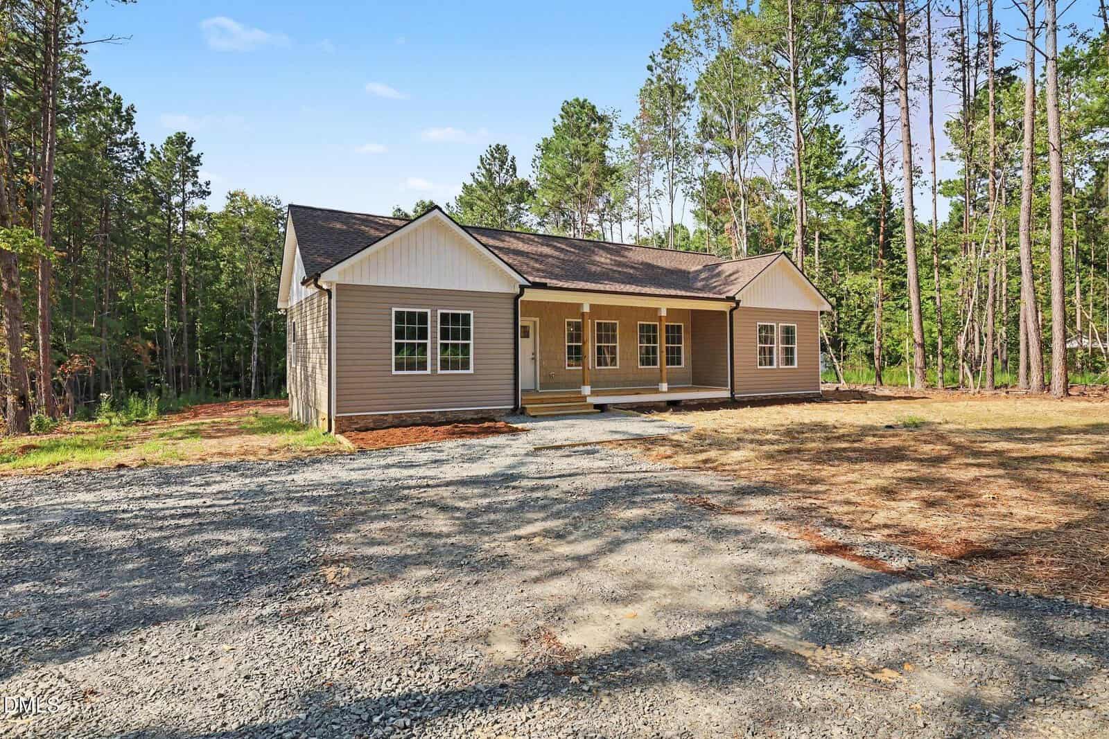 Single-story beige house with a covered front porch, white trim, and surrounded by tall pine trees. The yard is mostly dirt and gravel, with some grass and a forested background under a clear sky.