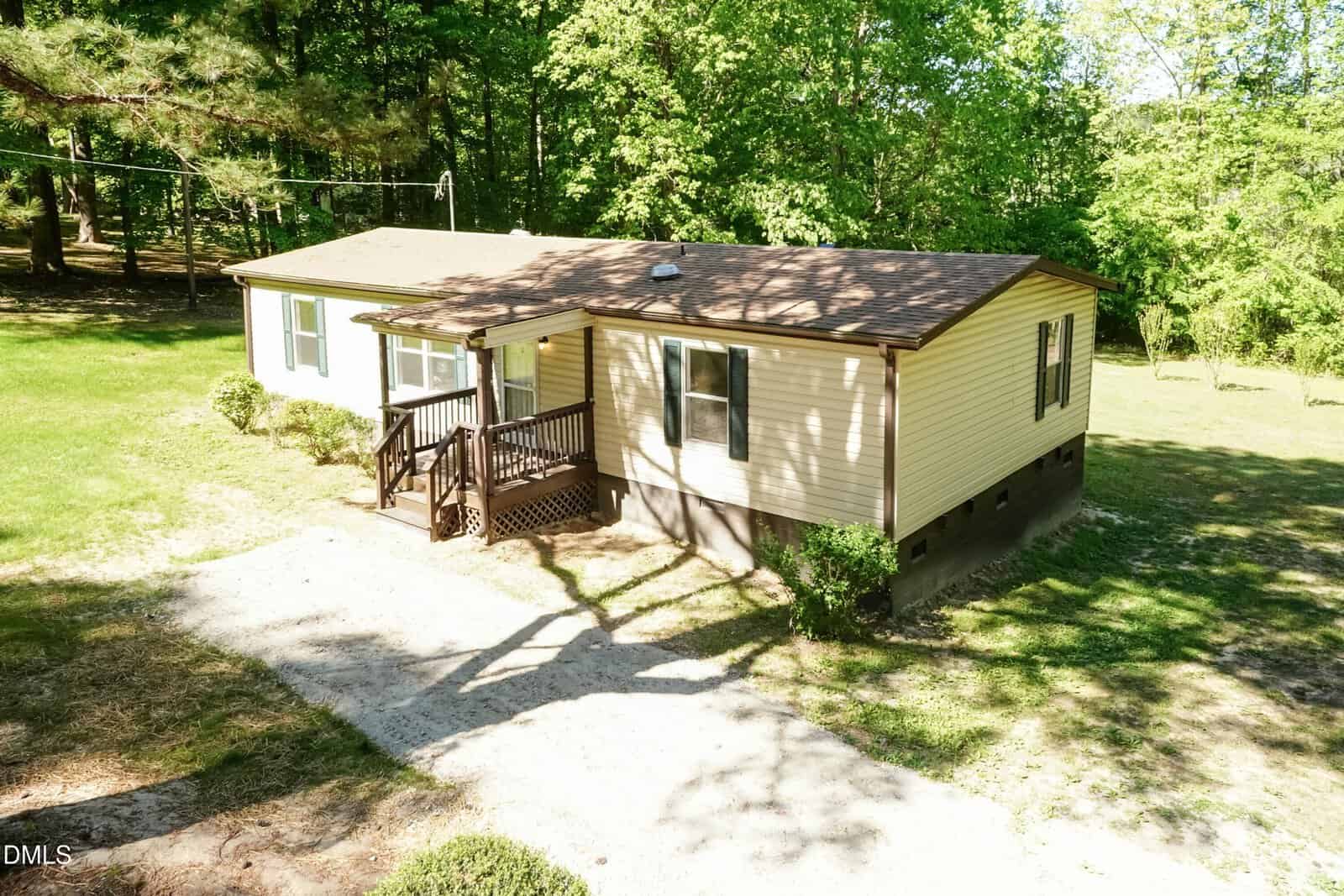 A tan single-story house with a brown roof and small front porch sits surrounded by green grass and trees, with a gravel driveway leading up to it.