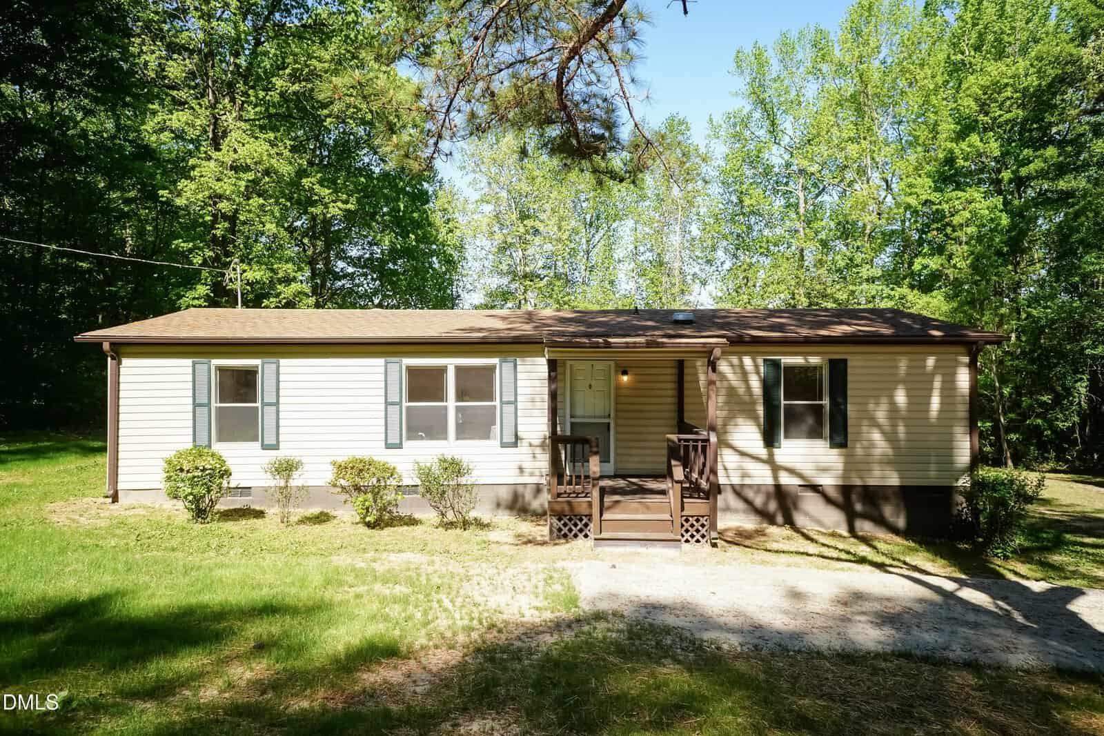 Single-story beige house with dark trim, small front porch, and green shutters, surrounded by trees and bushes on a sunny day. A dirt driveway leads up to the front steps of this charming home at 108 Edwards Drive in Louisburg.
