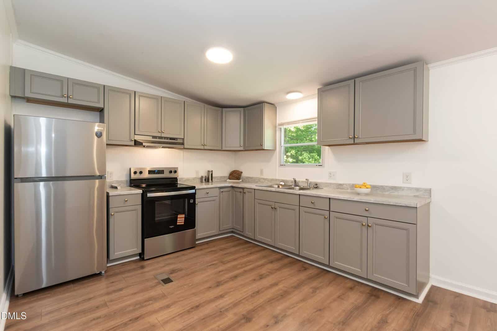 Modern kitchen with gray cabinets, stainless steel appliances, wood flooring, marble-patterned countertops, and a window above the sink letting in natural light. A bowl of lemons sits on the counter.