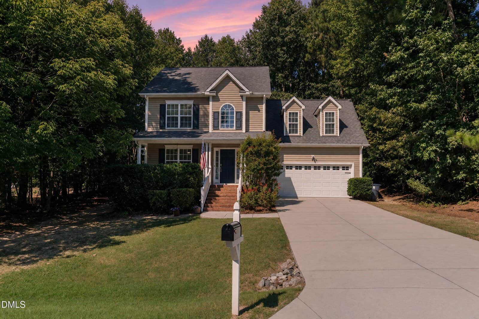 Two-story beige house with white trim, front porch, and two-car garage located at 85 Wilders Crossing Drive in Louisburg. Concrete driveway leads to mailbox; lush trees surround the home. Pink and purple sunset sky in the background.