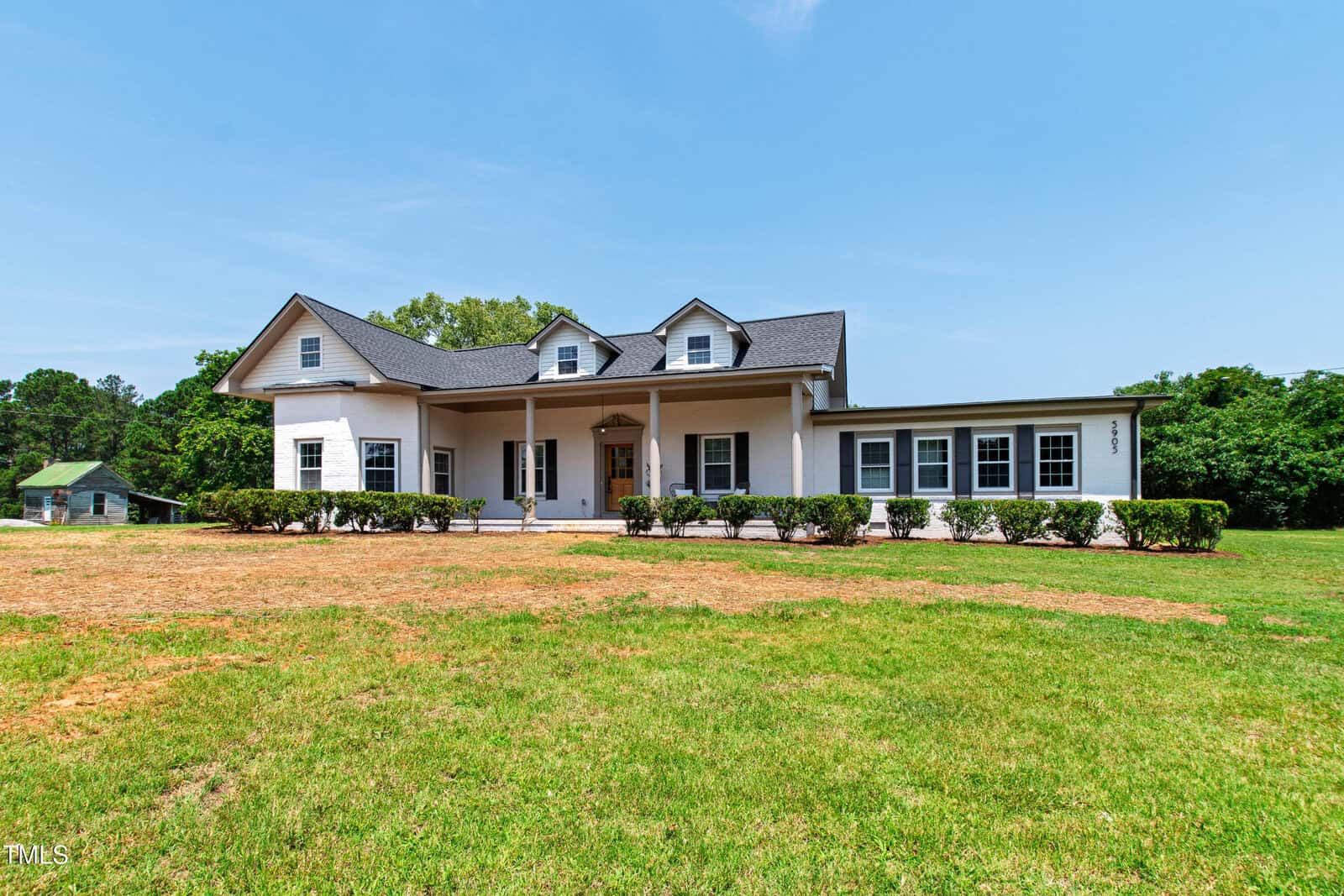 A large two-story white house with a front porch, multiple windows, and a grassy yard, set against a backdrop of green trees under a clear blue sky.