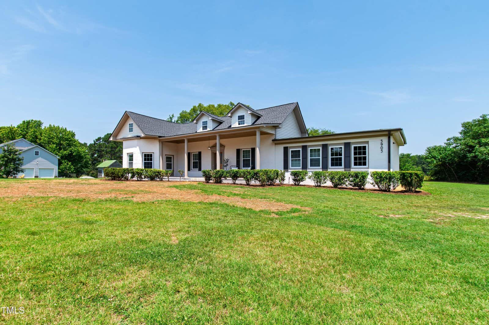 White modern farmhouse with dark roof, large front porch, dormer windows, and hedges along the front, set on a spacious grassy lawn with trees and a detached garage in the background under a clear sky.