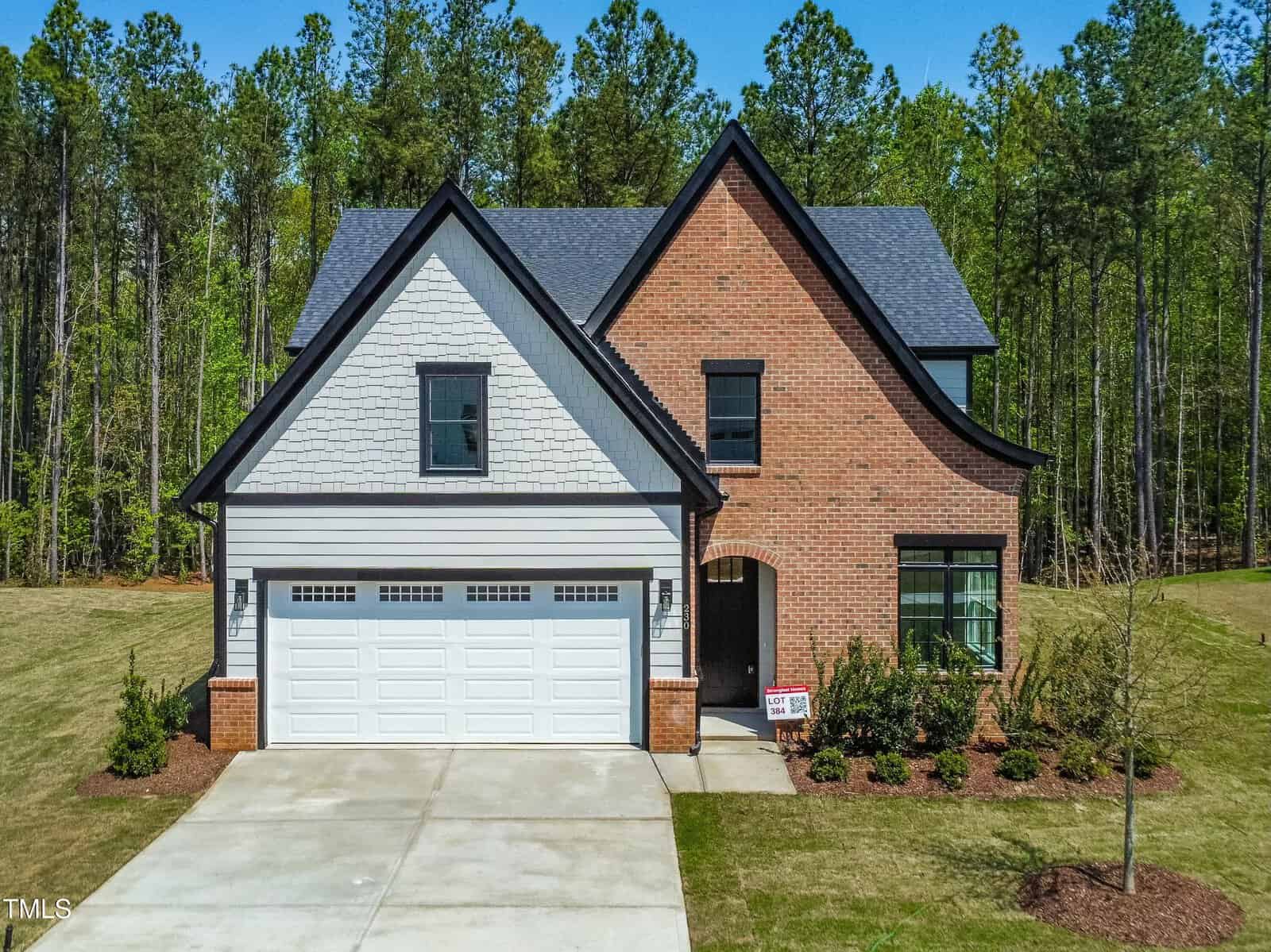 A two-story house with a brick and white exterior, black roof, and a double garage. It sits on a spacious lawn with trees in the background and a “For Sale” sign near the entrance.
