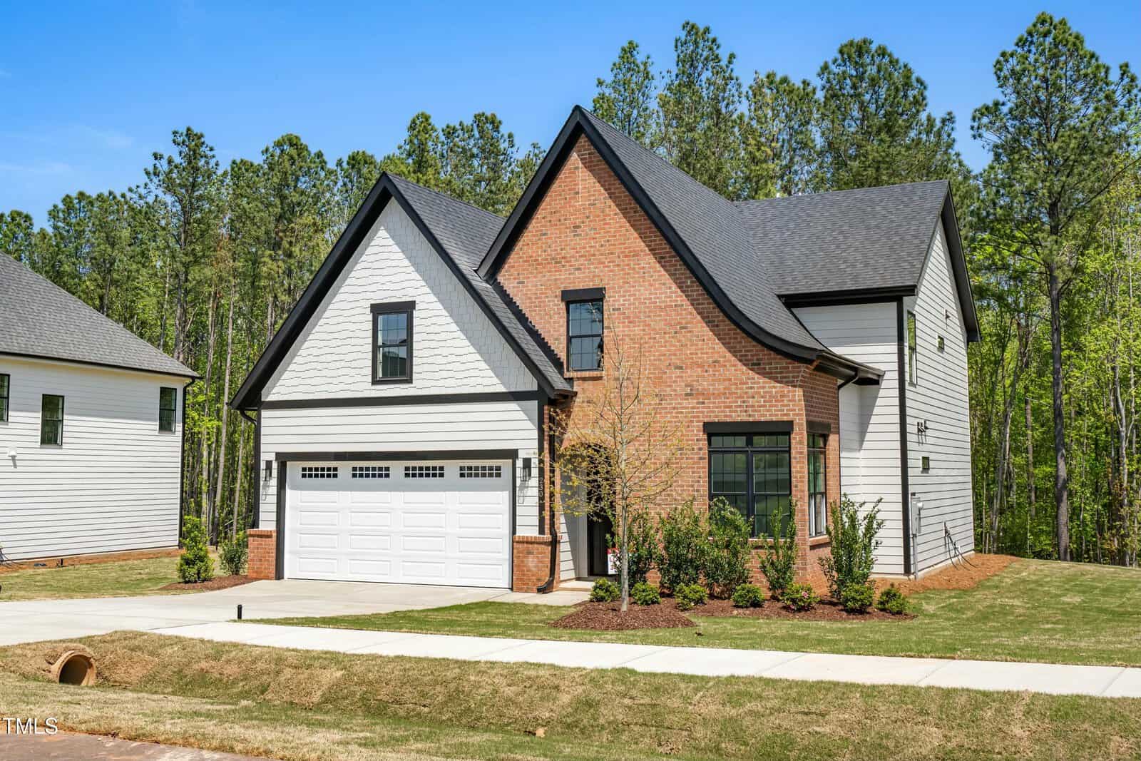Two-story suburban house with a brick and white siding exterior, attached two-car garage, small front yard, and surrounded by trees under a clear blue sky.