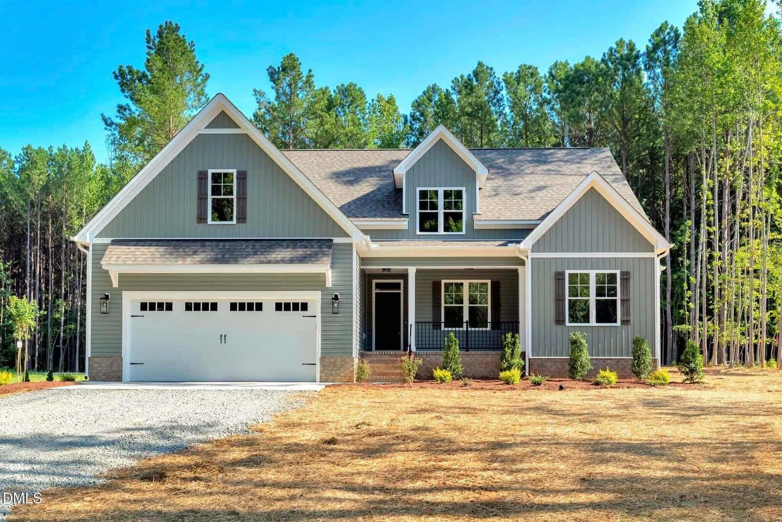 A modern gray house with white trim, a front porch, double garage, dormer windows, and manicured bushes sits gracefully on Lot 5 Watkins Wilkinson Road in Oxford, set against a backdrop of tall green trees and a clear blue sky.