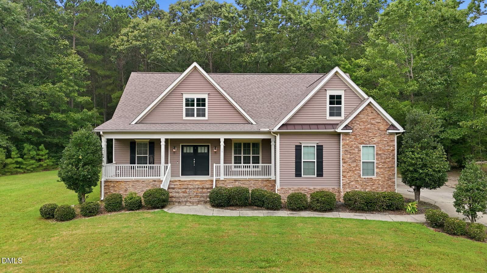 A two-story suburban house with a front porch, stone and siding exterior, black doors, white trim, and surrounded by green lawn, bushes, and trees.