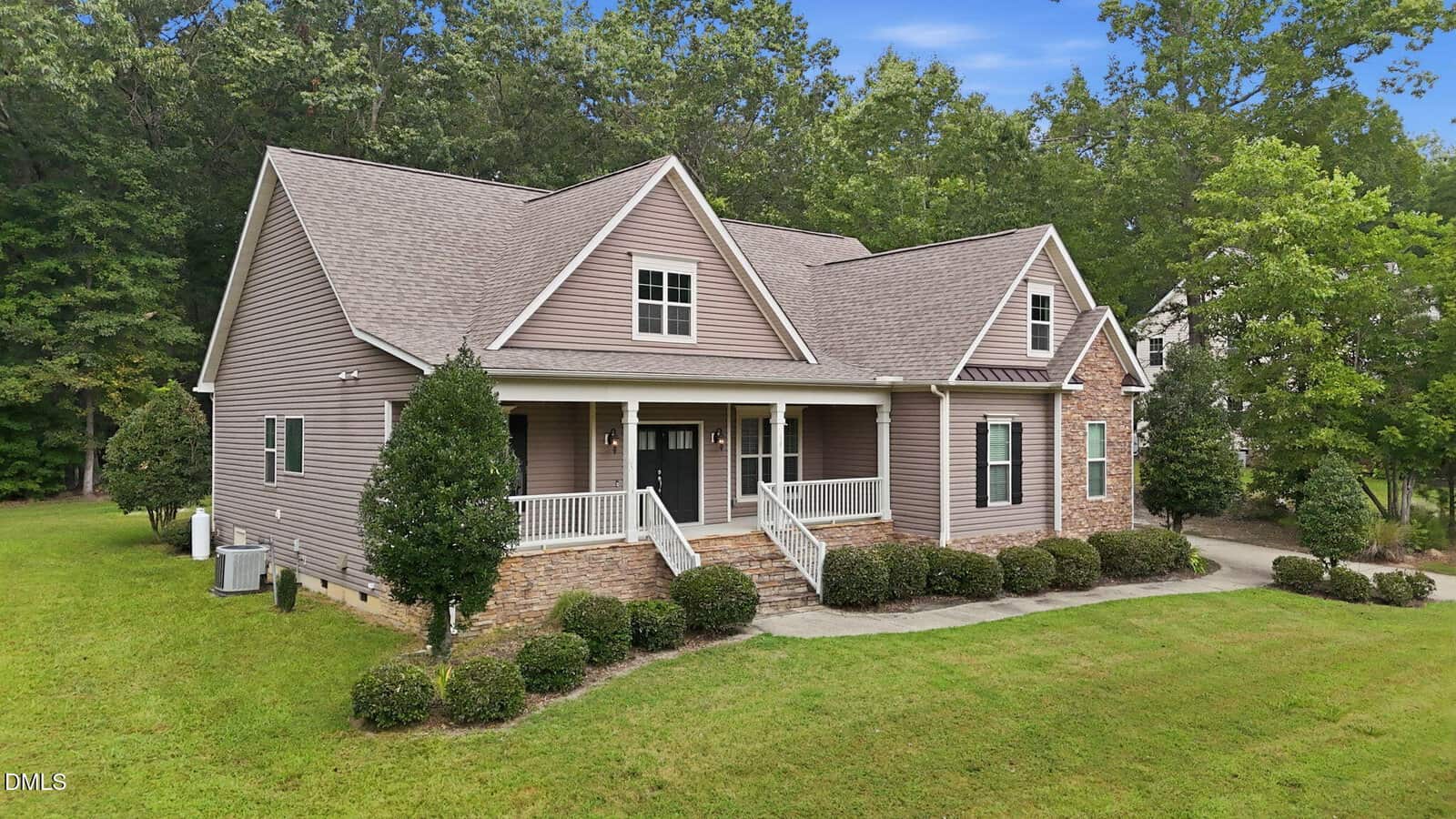 A beige two-story house with a gabled roof, white trim, front porch, brick accents, and manicured shrubs, set on a grassy lawn with trees in the background under a blue sky.
