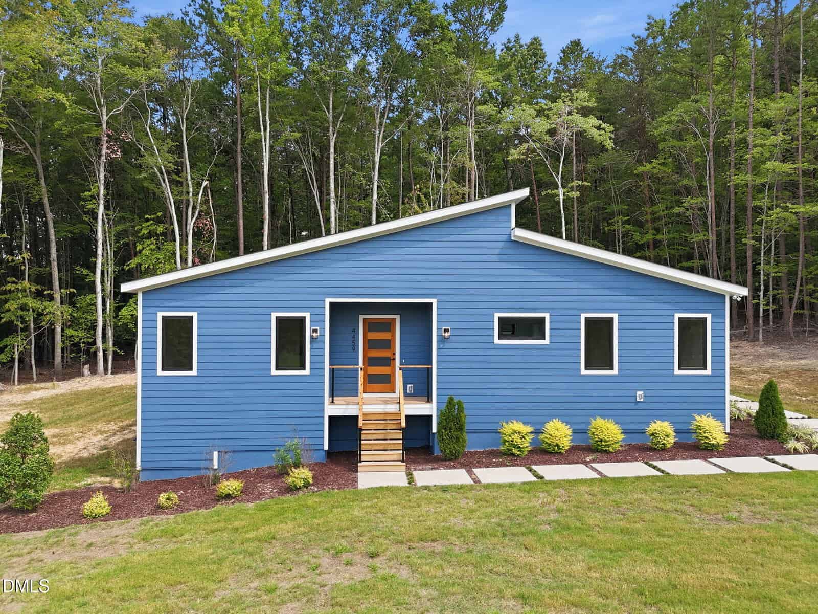 A modern blue house with a slanted roof, wooden front steps, and symmetrical windows, surrounded by green grass, shrubs, and trees in the background—perfectly situated on Lot 11 Poppy Mallow Lane in Oxford.