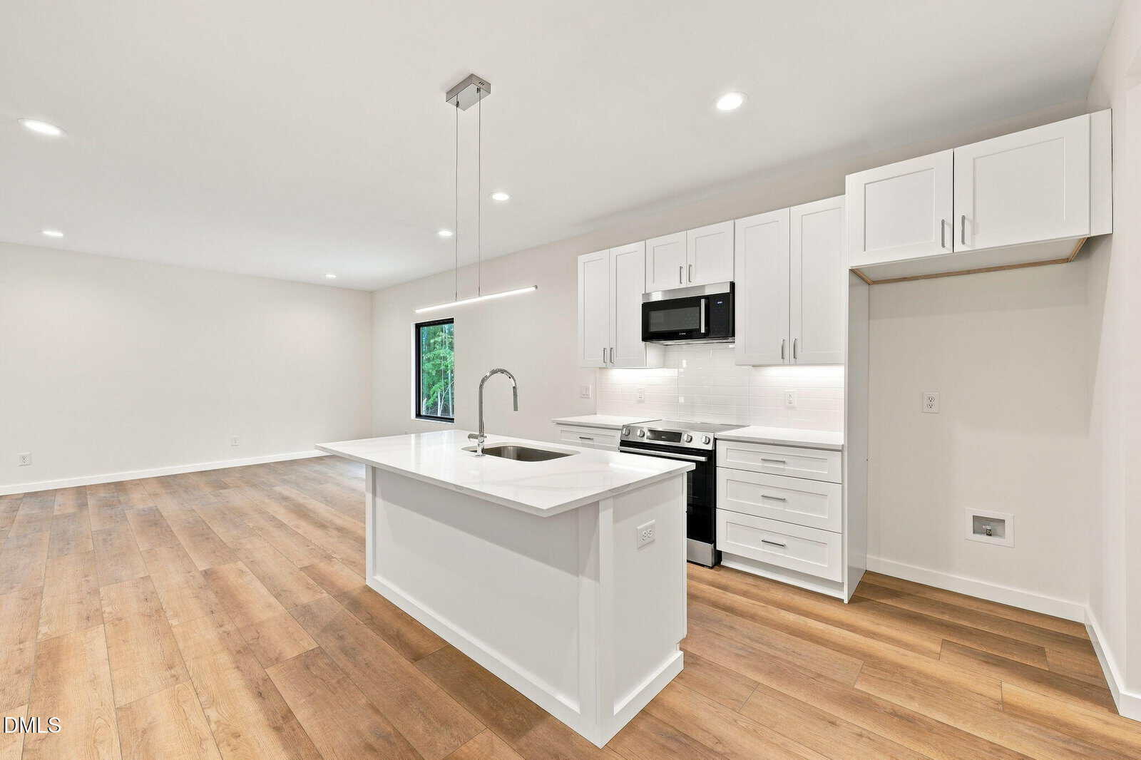 Modern kitchen with white cabinets, island with sink, stainless steel appliances, wood floors, and recessed lighting, open to a spacious living area with a large window.