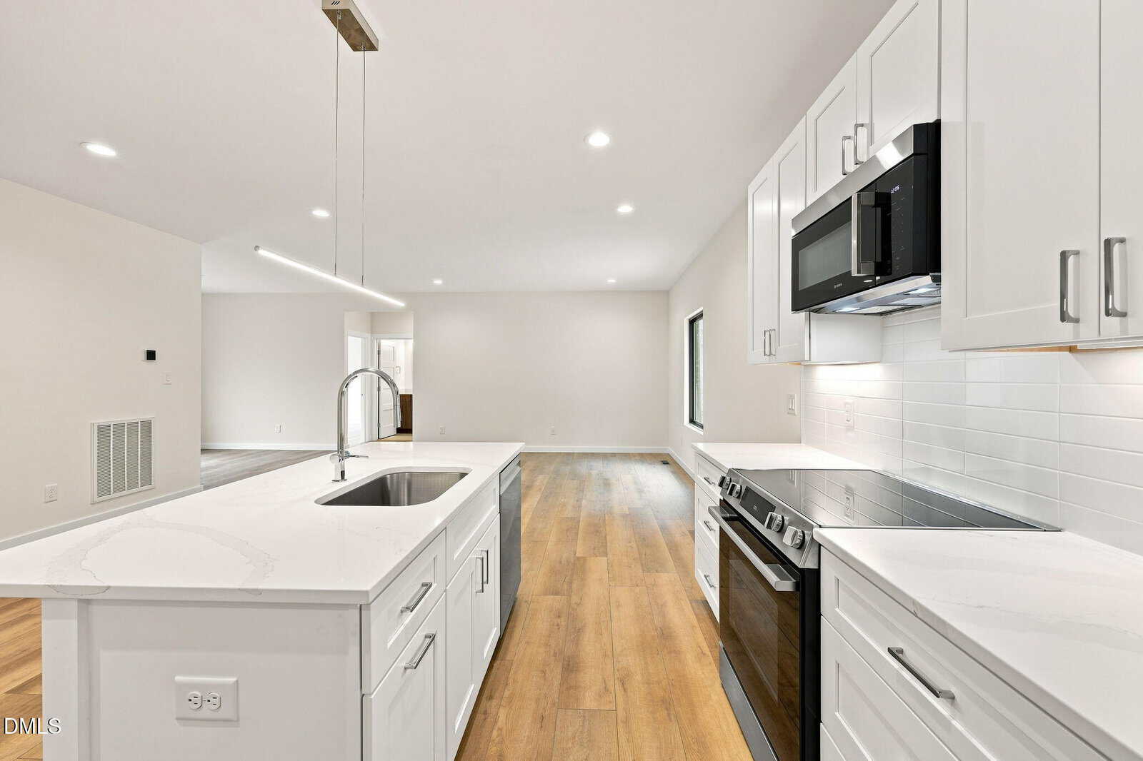 Modern kitchen with white cabinets, marble countertops, electric stove, built-in microwave, and sink island. The space has wood flooring, recessed lighting, and an open layout leading to a bright living area.