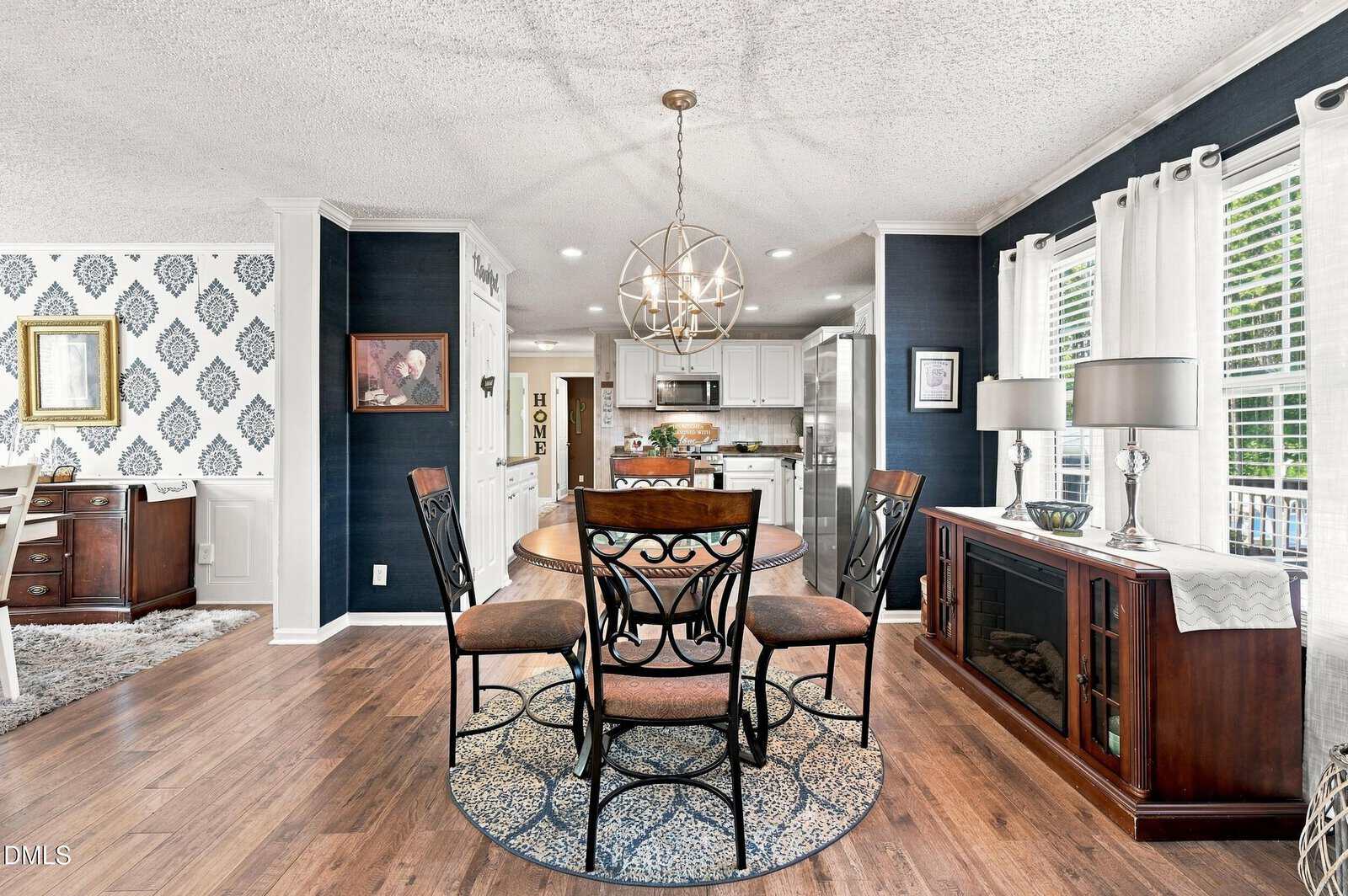 Modern dining area with round wooden table and four chairs, open to a white kitchen. Hardwood floors, dark accent walls, large windows with white curtains, and a decorative sideboard under two lamps.
