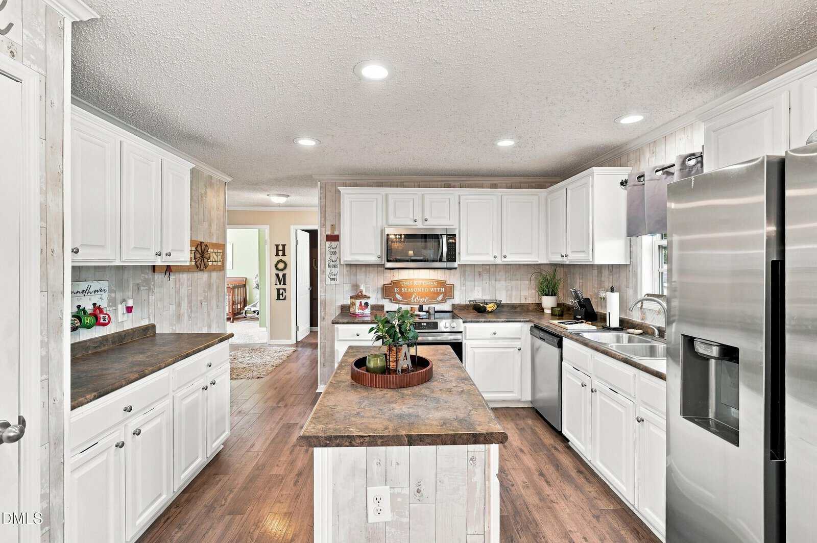 Bright kitchen with white cabinets, stainless steel appliances, a central island, wood floors, and decorative plants; a hallway and “HOME” sign are visible in the background.