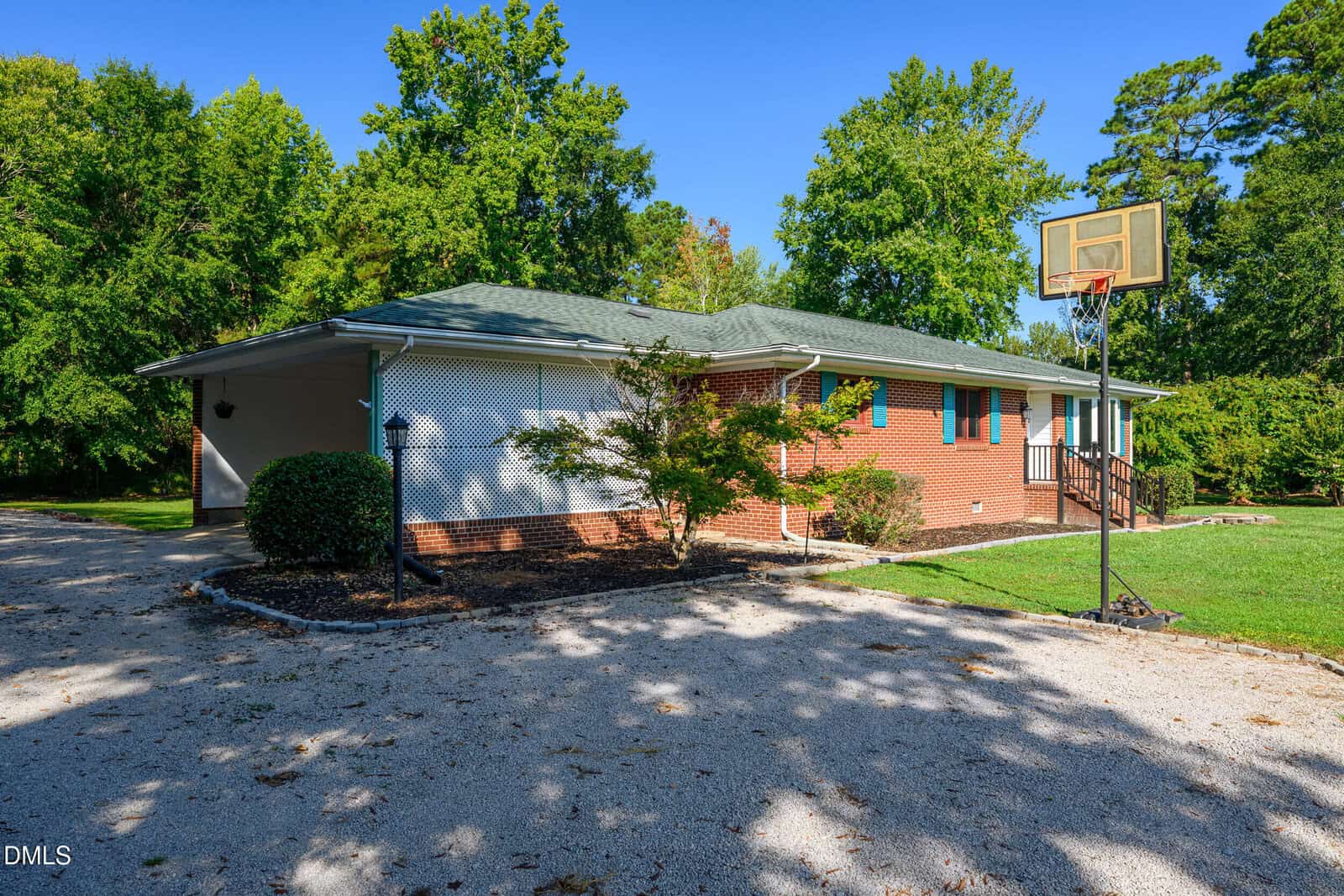 Single-story brick house with green roof, surrounded by trees. There’s a basketball hoop over a gravel driveway and a carport on the left side. The yard is grassy and well-maintained under a clear blue sky.