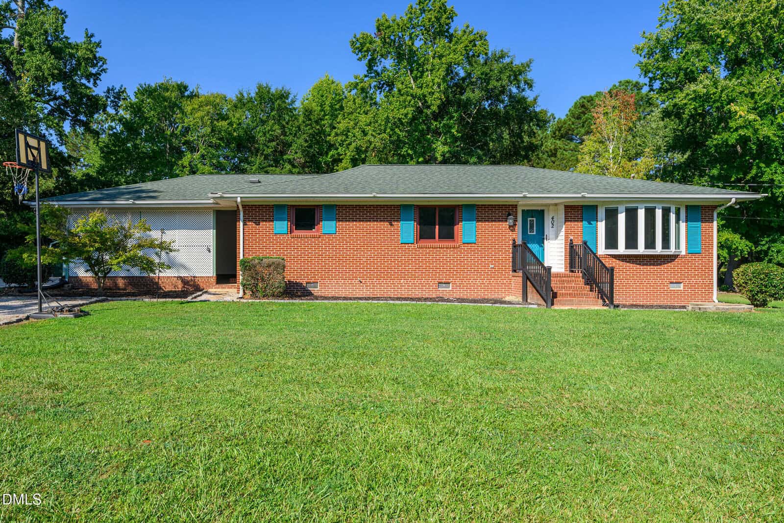 Single-story brick house with teal shutters, a two-car garage, front steps with black railing, and a large green lawn, surrounded by trees under a clear blue sky.