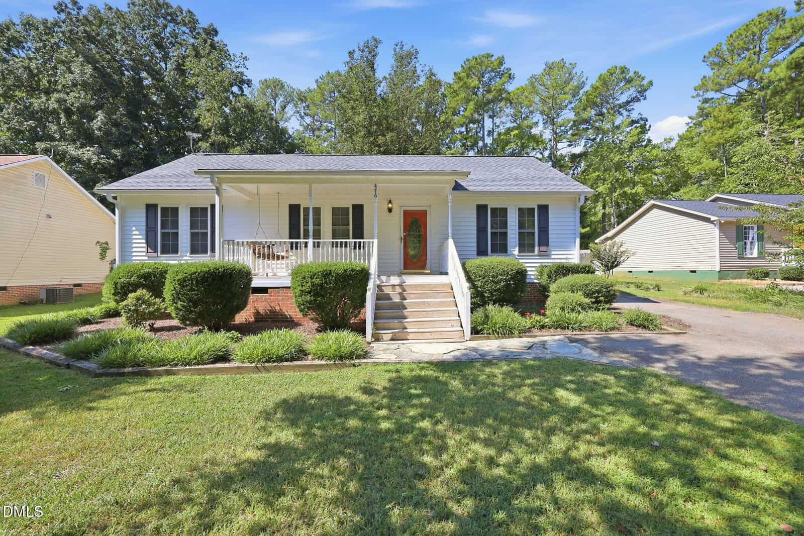 Single-story white house with a front porch, red door, porch swing, and trimmed bushes. Concrete steps lead to the entrance. The lawn is green and the driveway curves to the right. Trees are in the background.