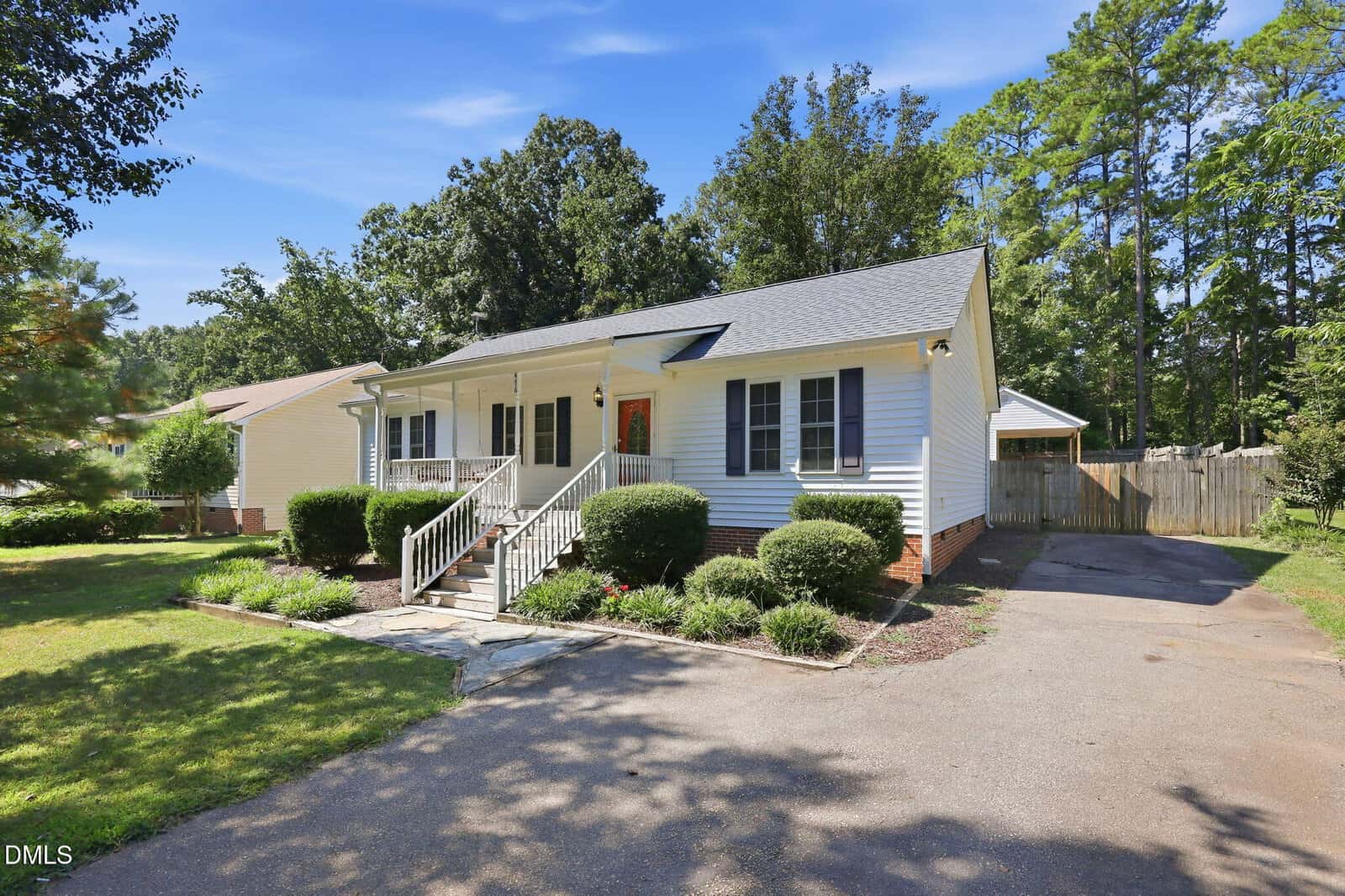 Single-story white house with gray shutters, a covered front porch, and neatly trimmed bushes. A driveway runs alongside the house, and a fenced backyard is visible. Tall trees surround the property under a clear blue sky.