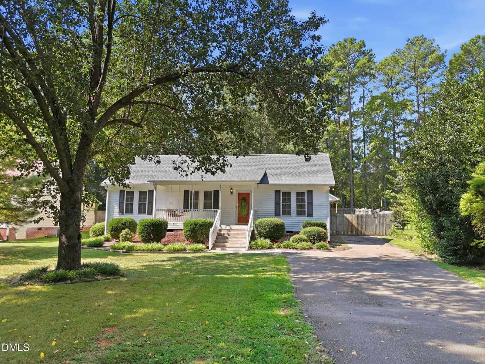 A small, white house with a front porch, red door, shrubs, and a large tree in the front yard sits at 476 Shawnee Drive in Louisburg. A driveway runs along the right side, and tall pine trees stand in the background.
