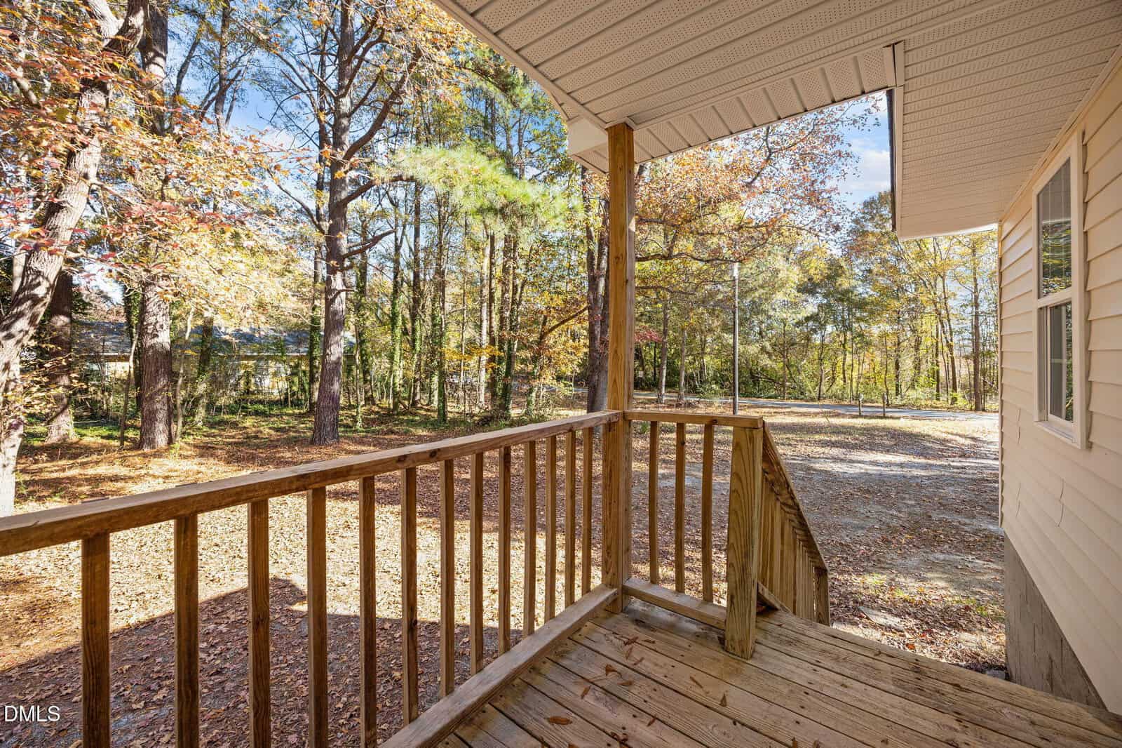 View from a wooden porch with railings, overlooking a gravel driveway and a yard filled with tall trees. The ground is covered with fallen autumn leaves under a clear sky.