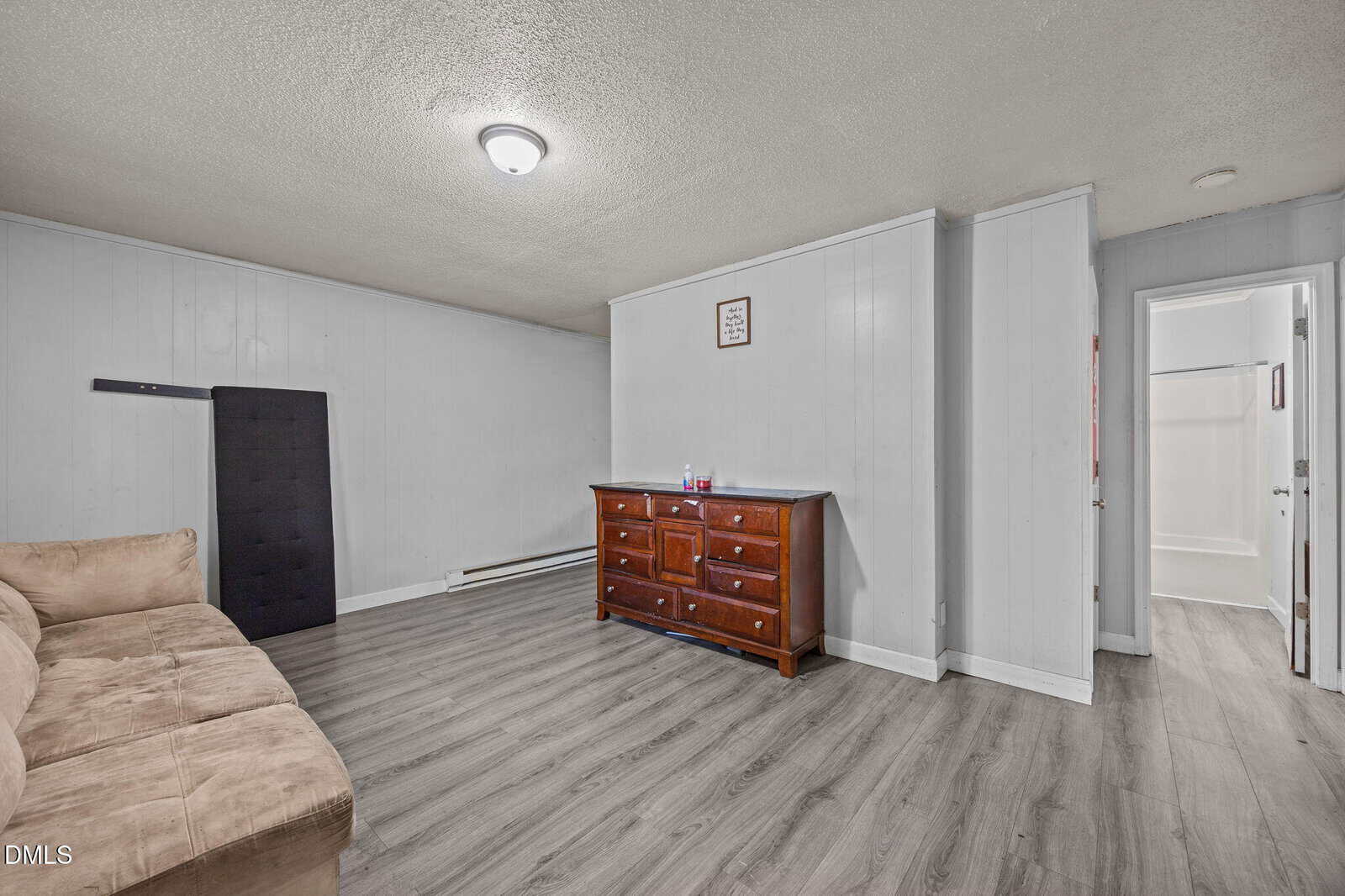 A simple room with gray wood floors, a tan couch, a wooden dresser, a wall-mounted panel, and an open doorway leading to a bathroom with a white tub and shower.