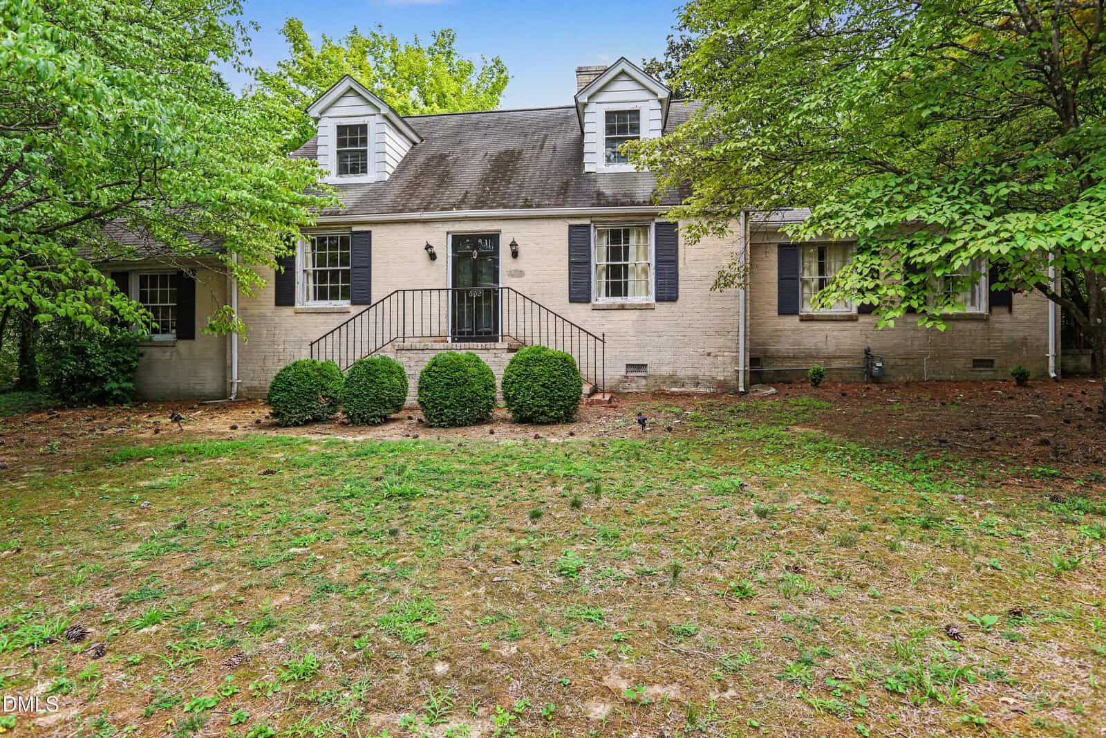 A brick house at 602 Williamsboro Street in Oxford, with a front staircase, dark shutters, dormer windows, and bushes along the front, is surrounded by trees and shown on a patchy, grassy yard.