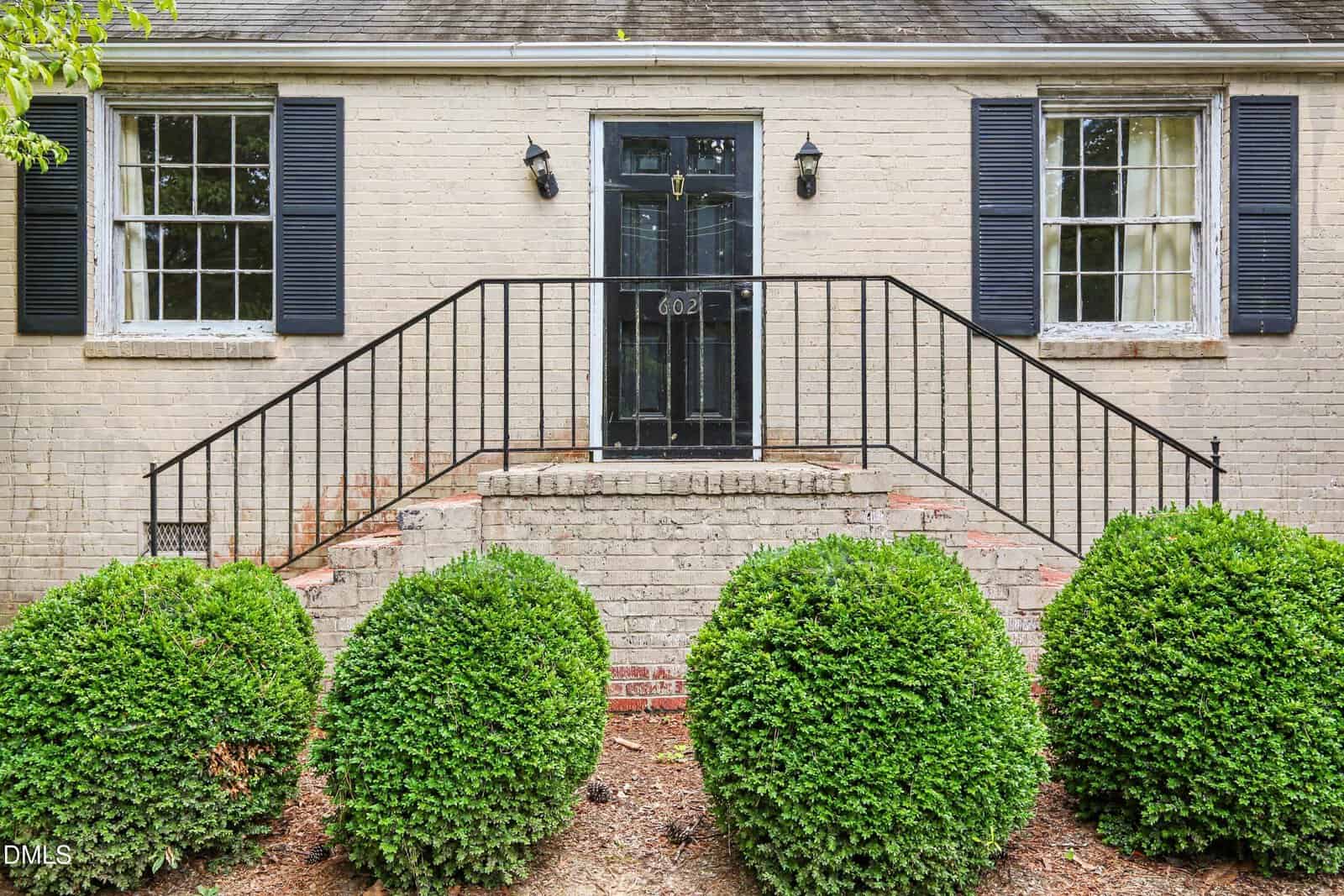 A brick house with black shutters and door, metal railing on stairs, and five trimmed bushes along the front. House number 602 is visible on the door.