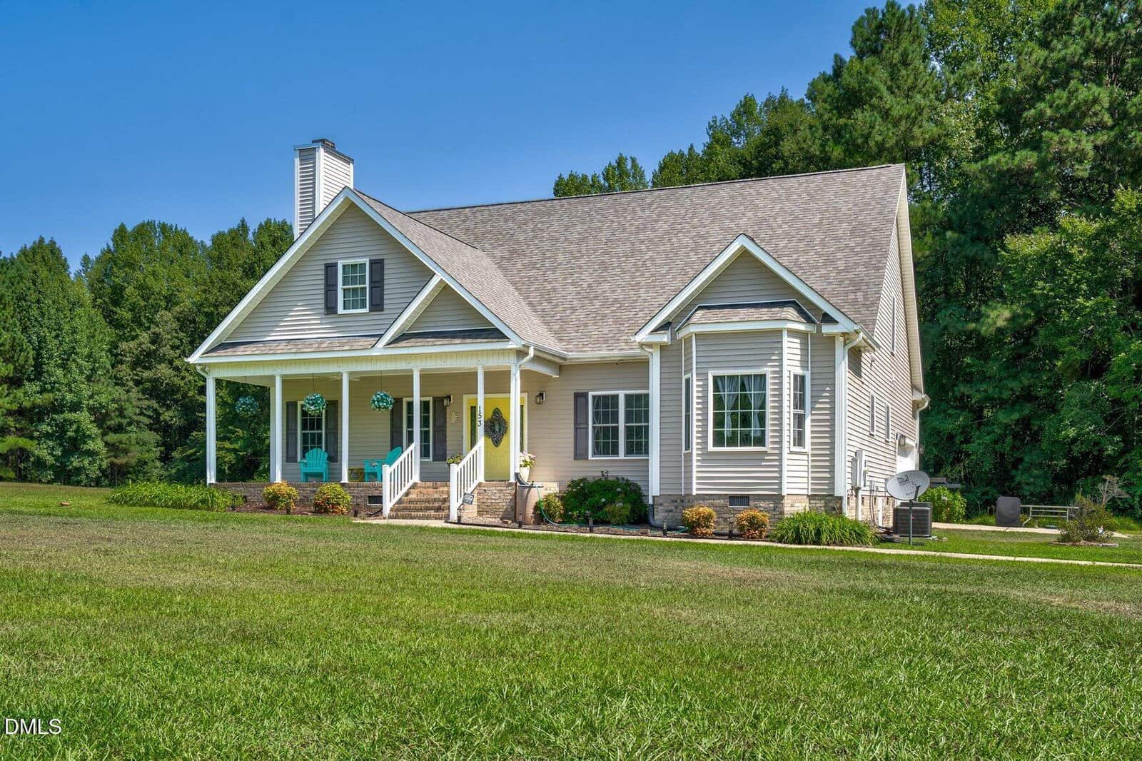 A light-colored house with a covered front porch, gabled roof, and steps leading to the entrance, surrounded by green grass and trees in the background on a sunny day.