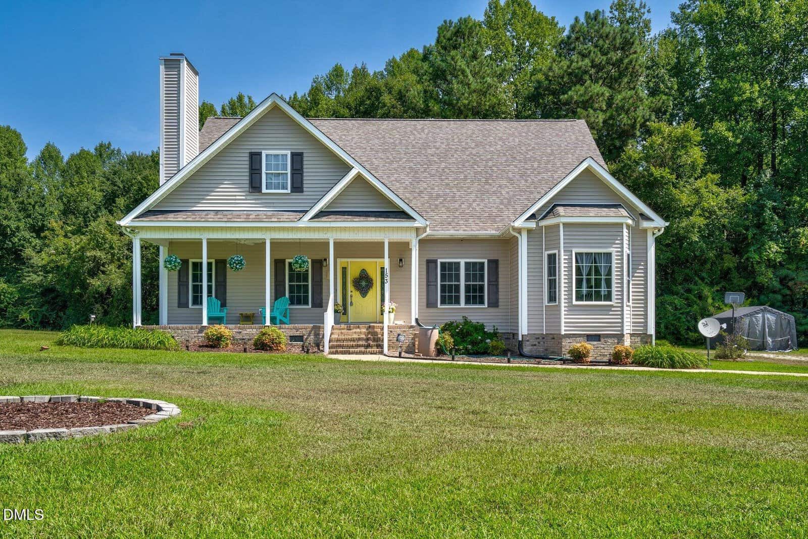 A white, single-story house at 153 Meadowbrook Lane in Spring Hope features a front porch with blue chairs, hanging plants, and a well-kept lawn, all surrounded by trees under a clear blue sky.