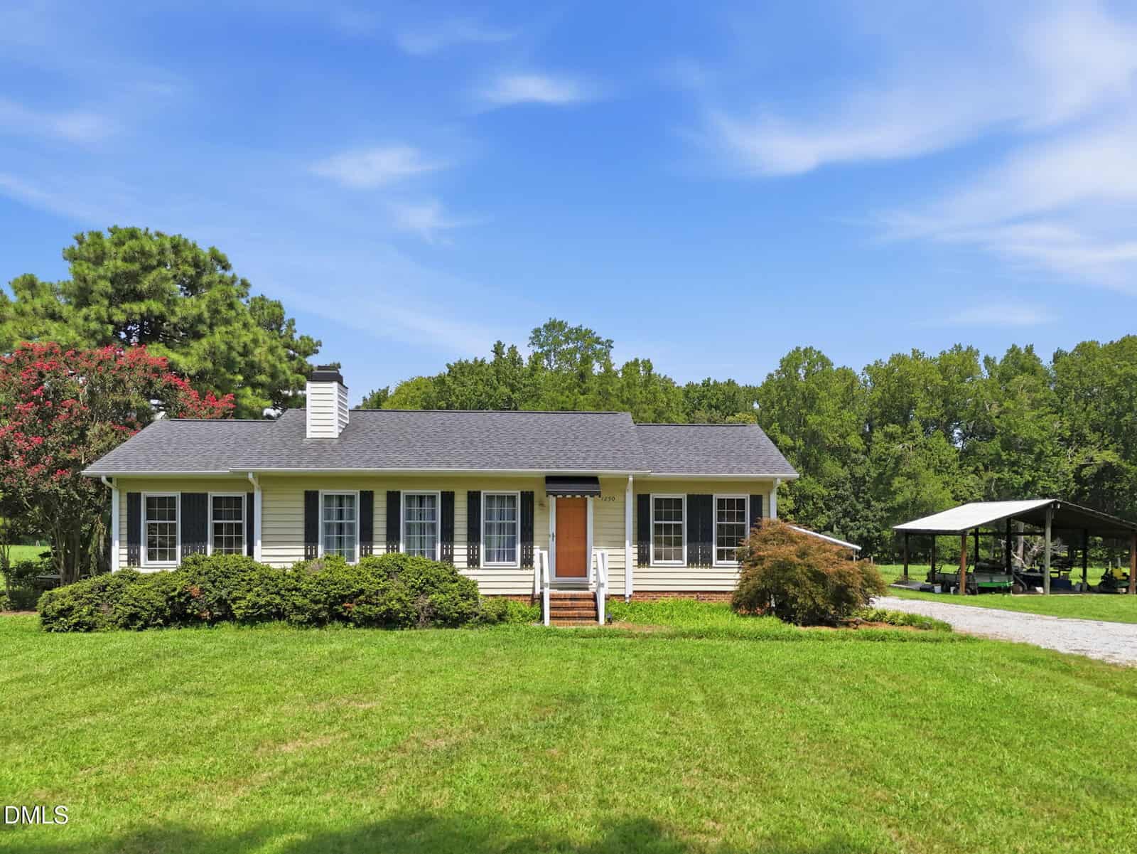 Charming single-story house at 1290 Spring Valley Road, Henderson, featuring classic white siding, black shutters, and a welcoming front porch. The home is surrounded by a green lawn and mature trees. To the right, a gravel driveway leads to a convenient carport. Overhead, a blue sky with light clouds completes this peaceful setting.