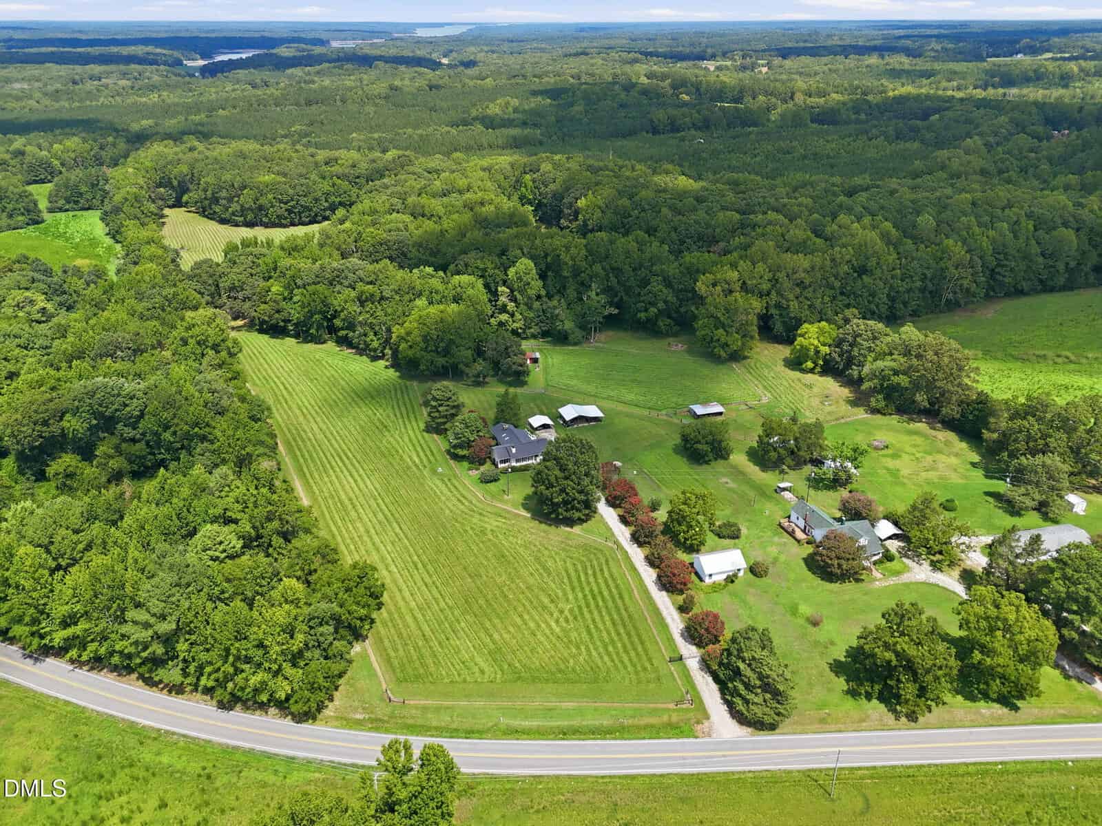 Aerial view of a rural property with several houses, barns, manicured lawns, and surrounding forest, bordered by a road and distant hills under a partly cloudy sky.