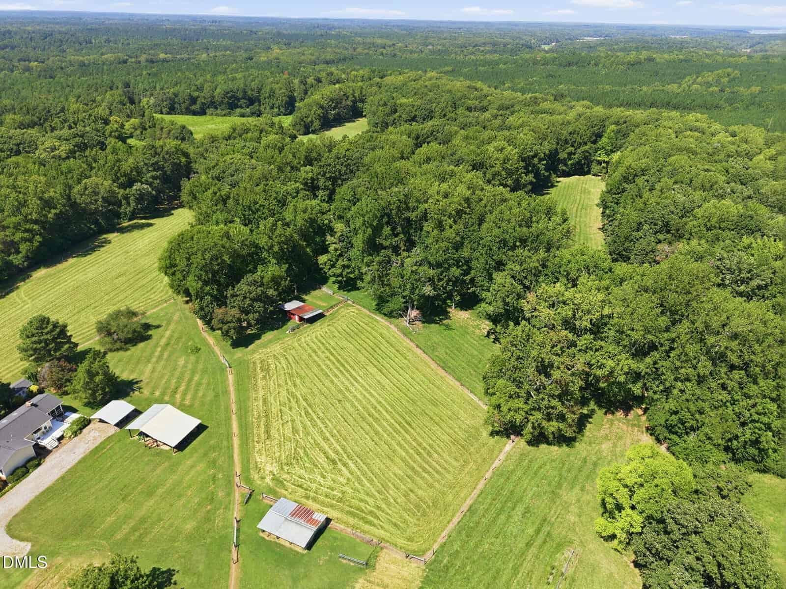 Aerial view of Lot 2 Spring Valley Road, Henderson, showcasing mowed fields, several buildings, fenced pastures, and dense green forests surrounding the area under a partly cloudy sky.
