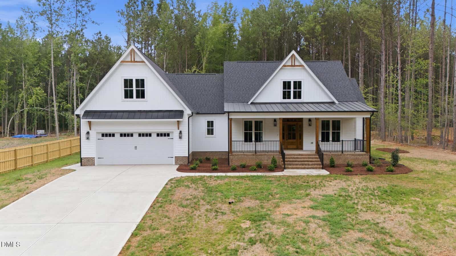 A modern white house with a black roof, double garage, front porch, and neatly landscaped yard sits in front of a forest with tall trees at 1157 Shonele Lane. This stunning home blends seamlessly into its natural surroundings with stem landscaping that enhances its curb appeal.