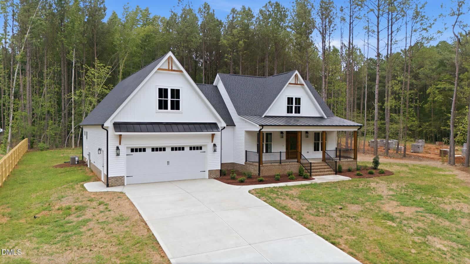 A modern white house with black trim, a two-car garage, front porch, and large driveway, surrounded by grass and tall trees in a wooded area.