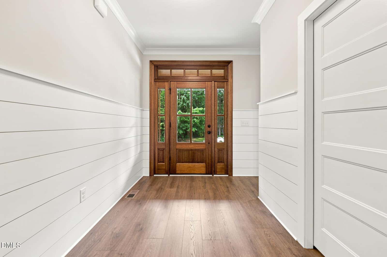 Bright hallway with white shiplap walls, wood flooring, and a wooden front door with glass panels. Green trees are visible through the door’s windows.