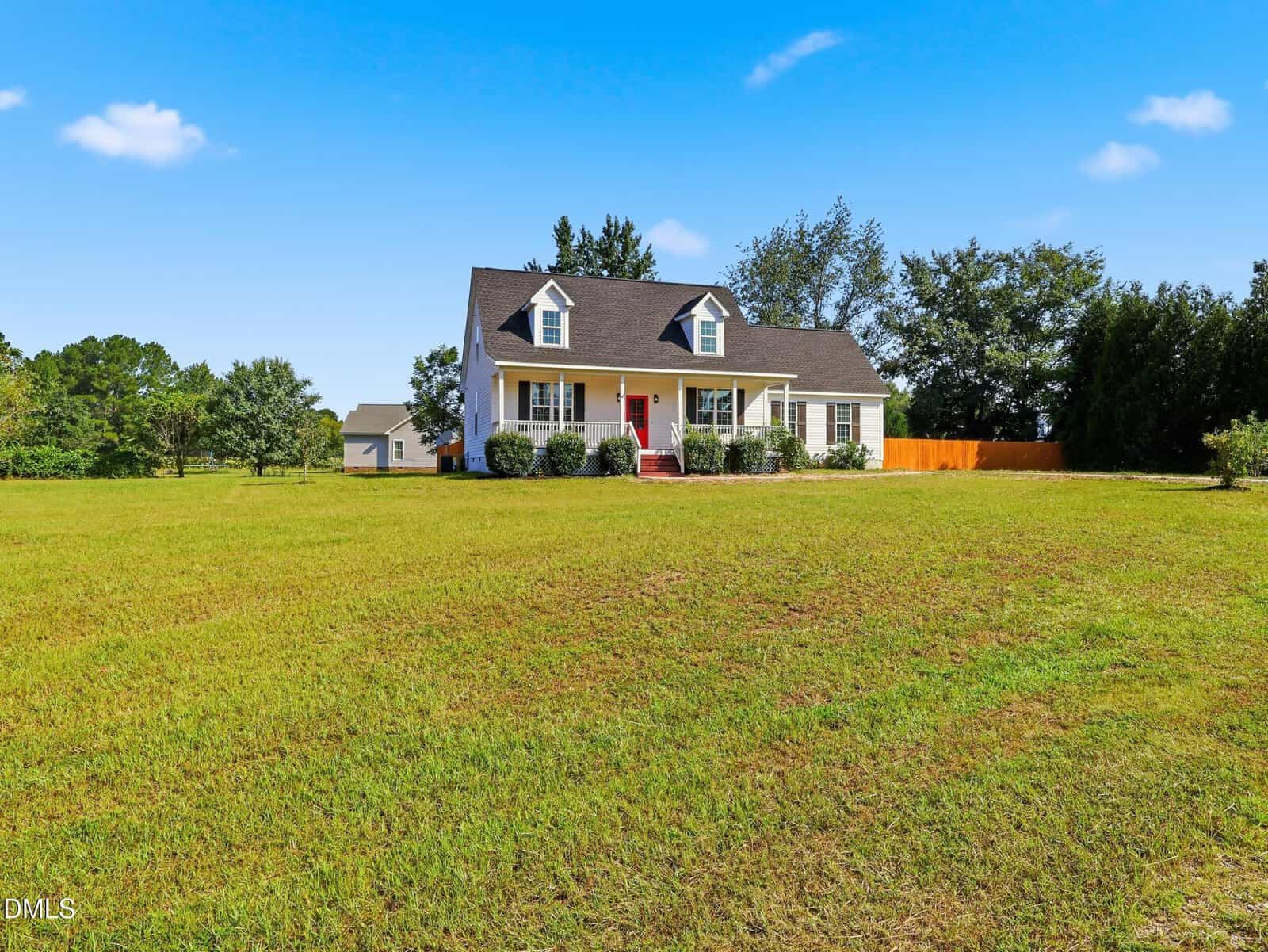 A white two-story house with dormer windows and a red front door sits on a large, open grassy lawn under a blue sky with a few clouds. Trees and a wooden fence are in the background.