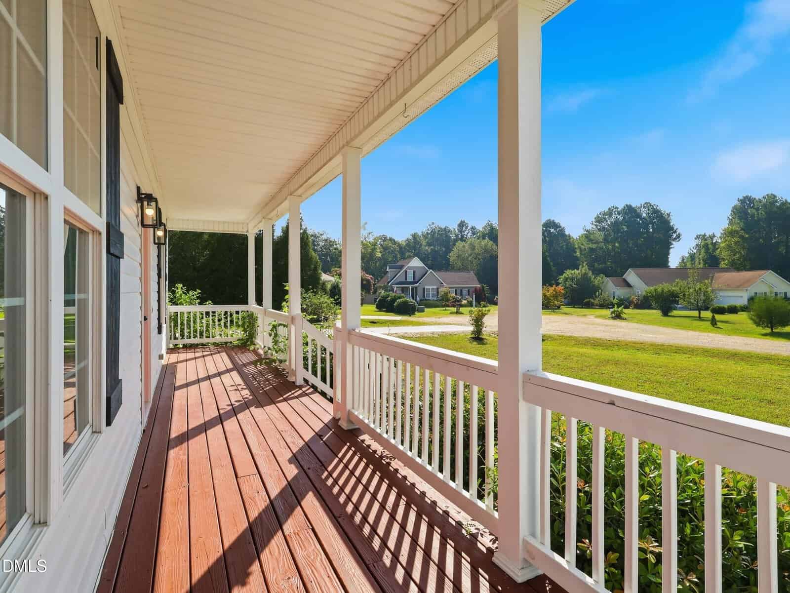 A wide view from a white wooden porch with railings and a polished wood floor, overlooking a green lawn, trees, and neighboring houses under a clear blue sky.